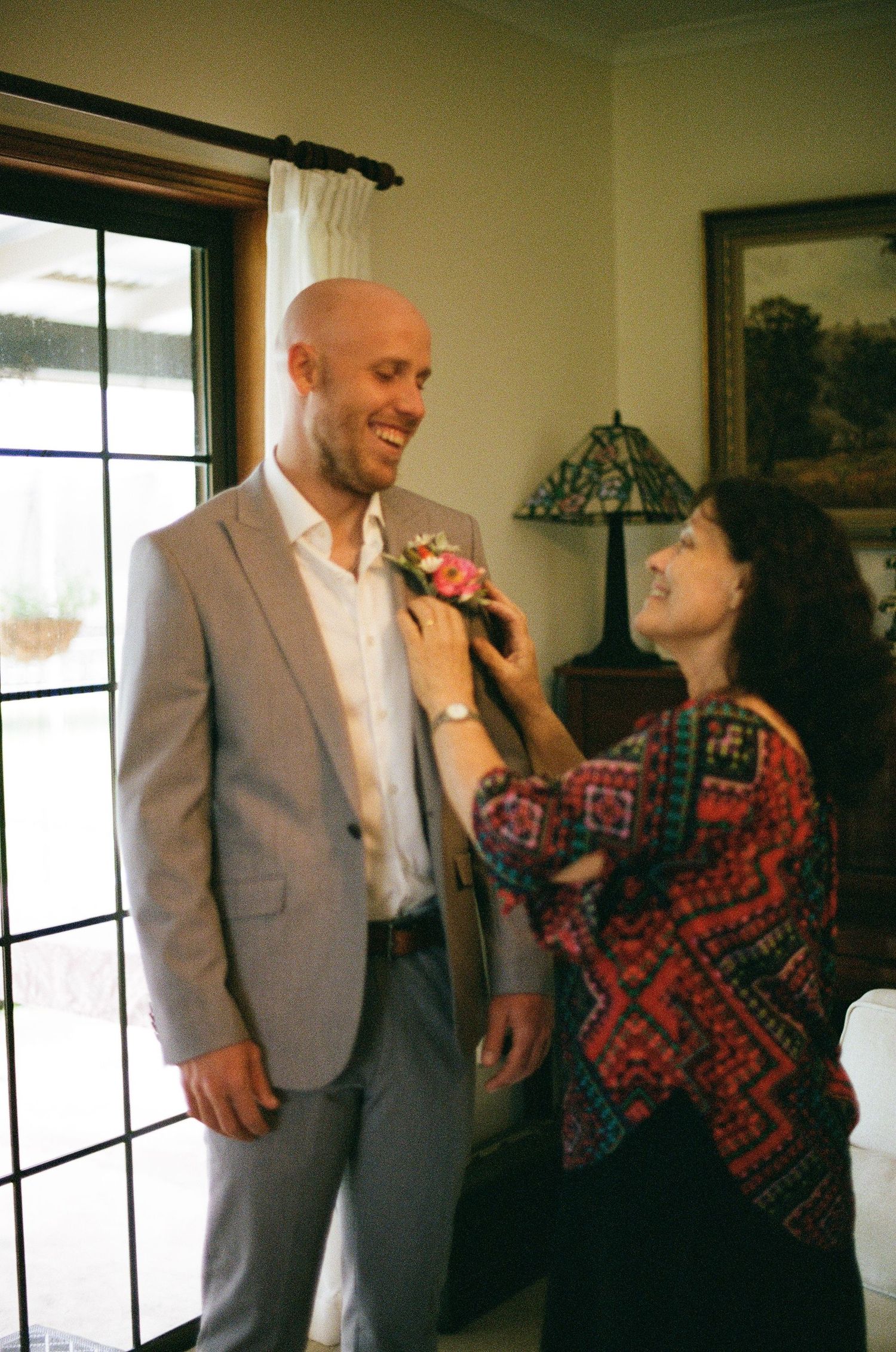 Smiling bald groom in gray suit gets boutonniere pinned by woman in colorful patterned jacket indoors.