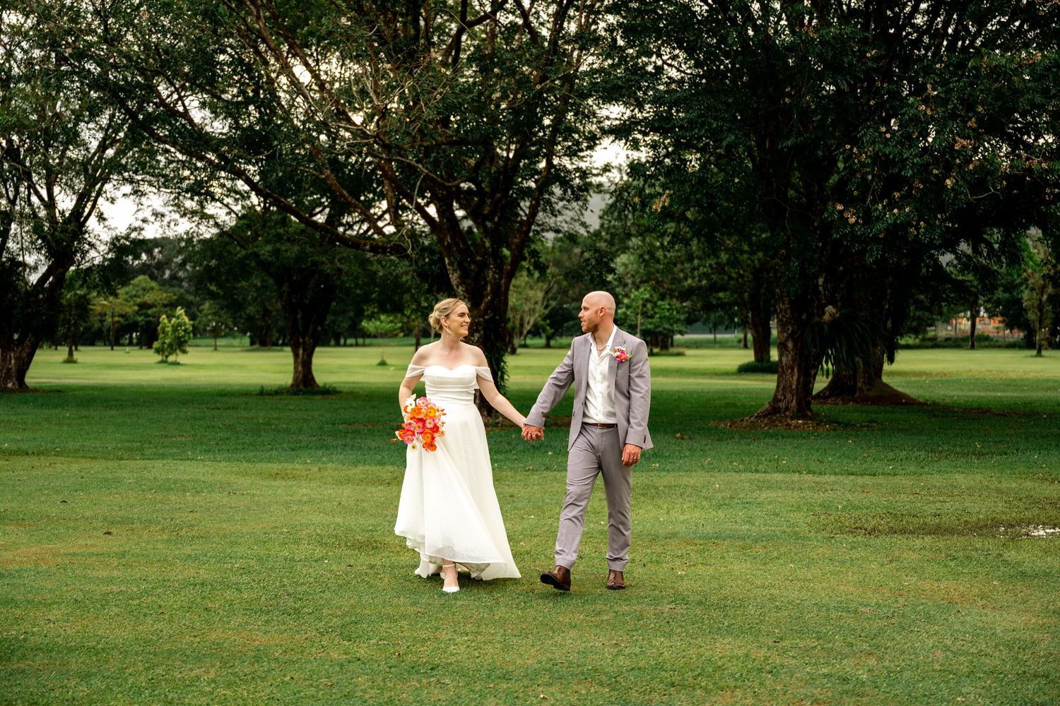 Bride and groom holding hands walking across green lawn surrounded by trees, bride carrying vibrant orange bouquet.