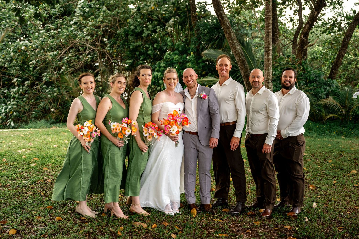 Wedding party group photo outdoors with bride in white, bridesmaids in green dresses holding vibrant orange bouquets, and groomsmen.