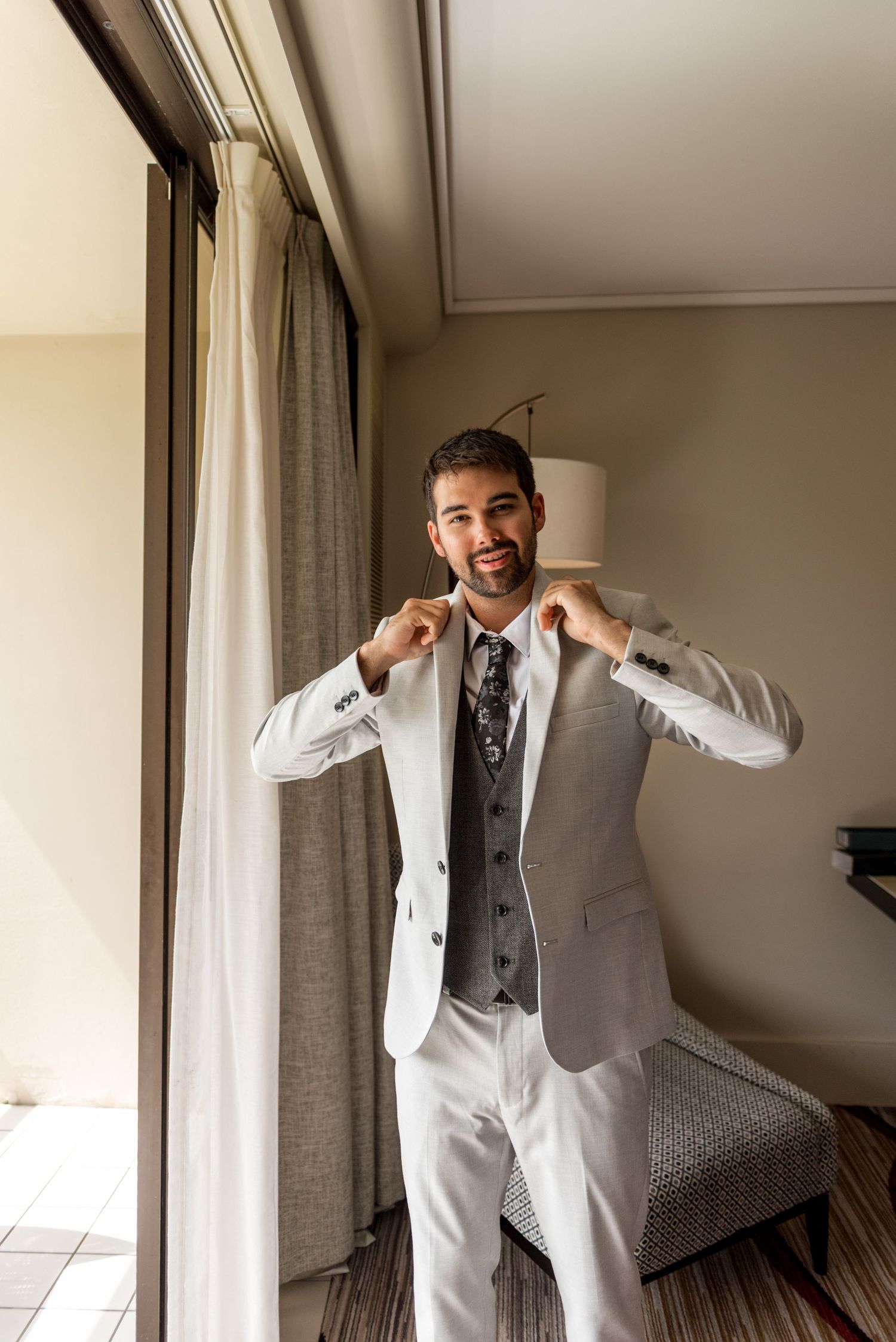 Smiling groom in light gray suit adjusting his collar by a hotel window on his wedding day.