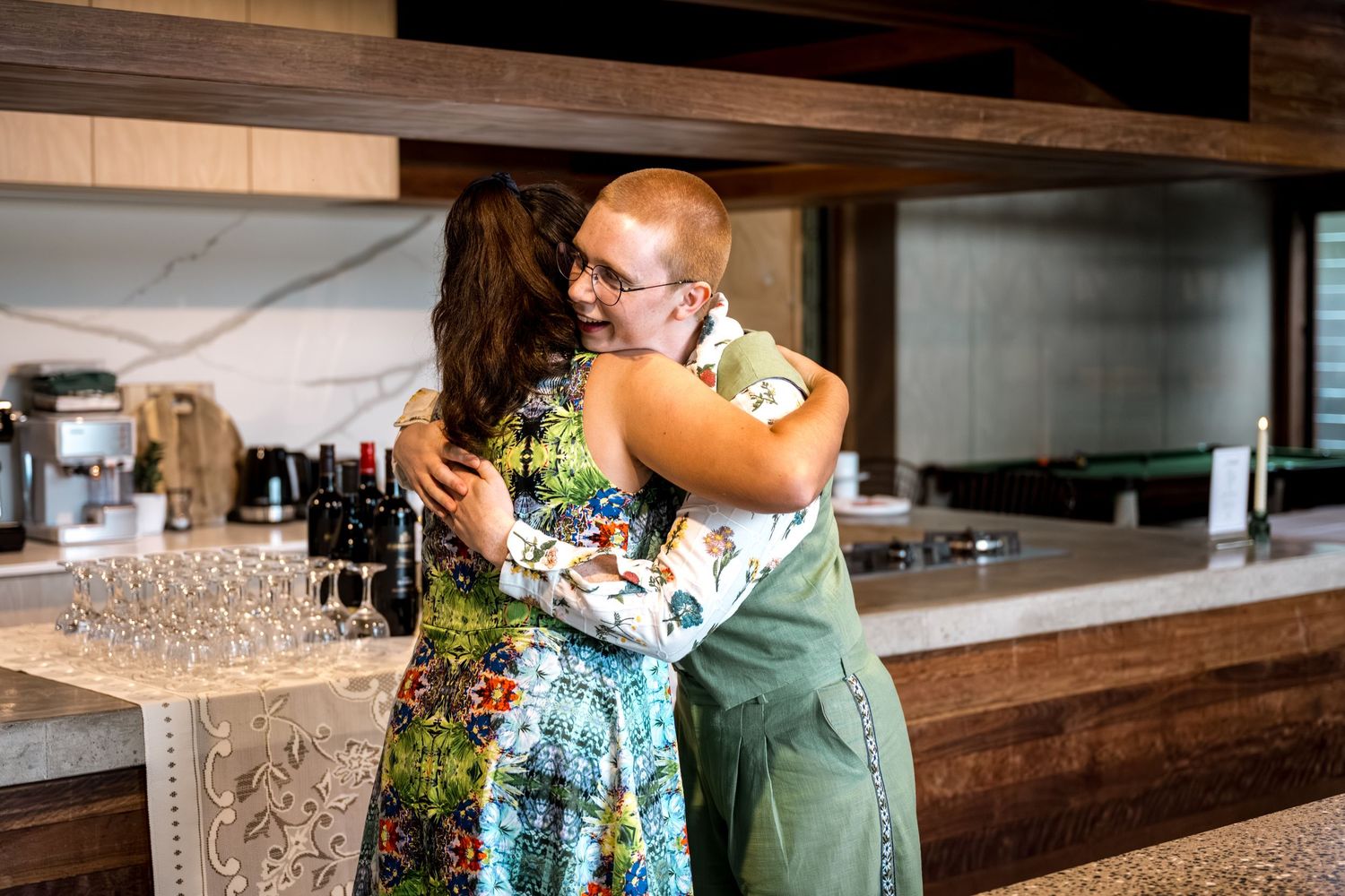 Two guests share a warm hug near the bar at an indoor wedding reception.