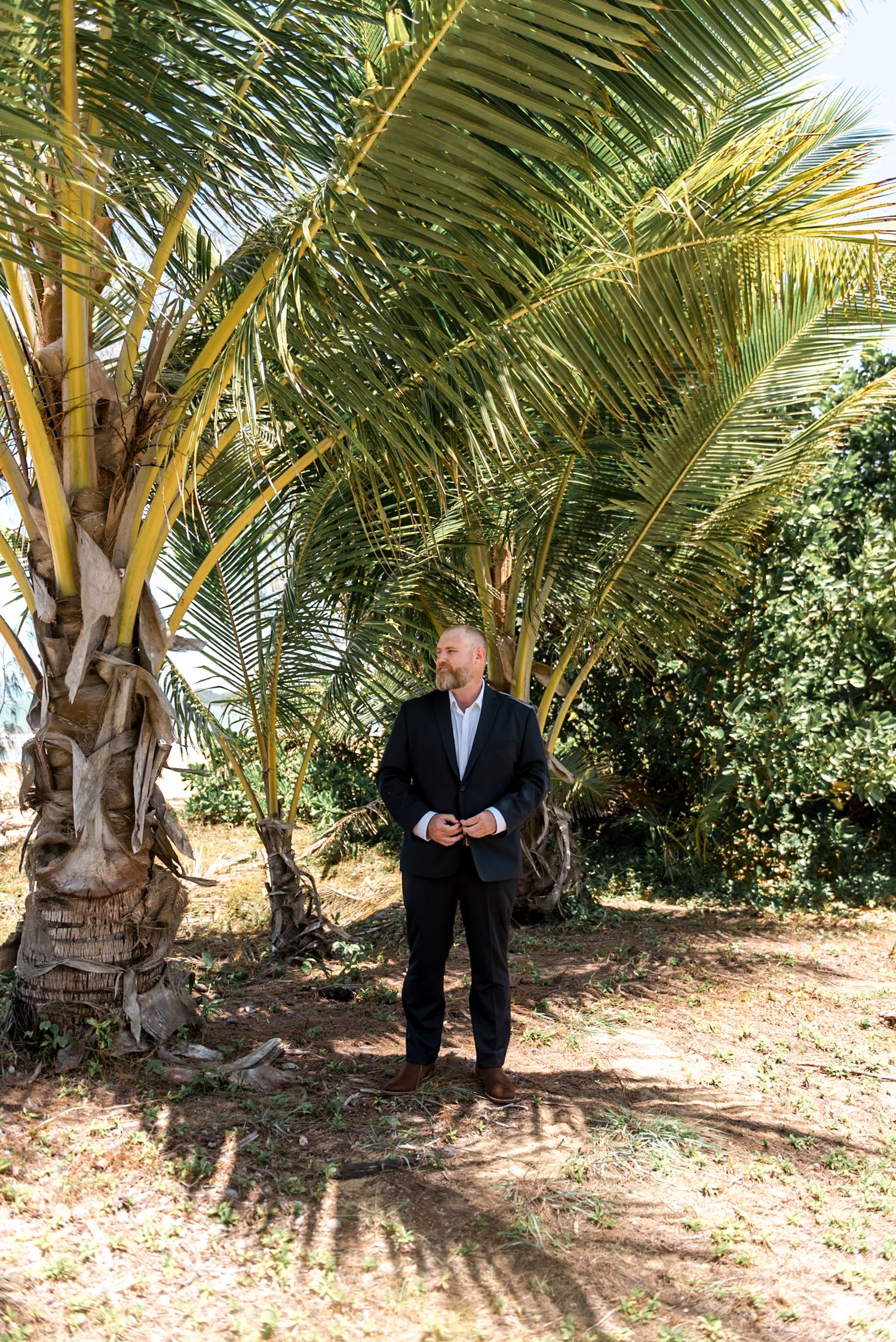 Dapper man in dark suit standing confidently beneath tall tropical palm trees in bright sunlight.