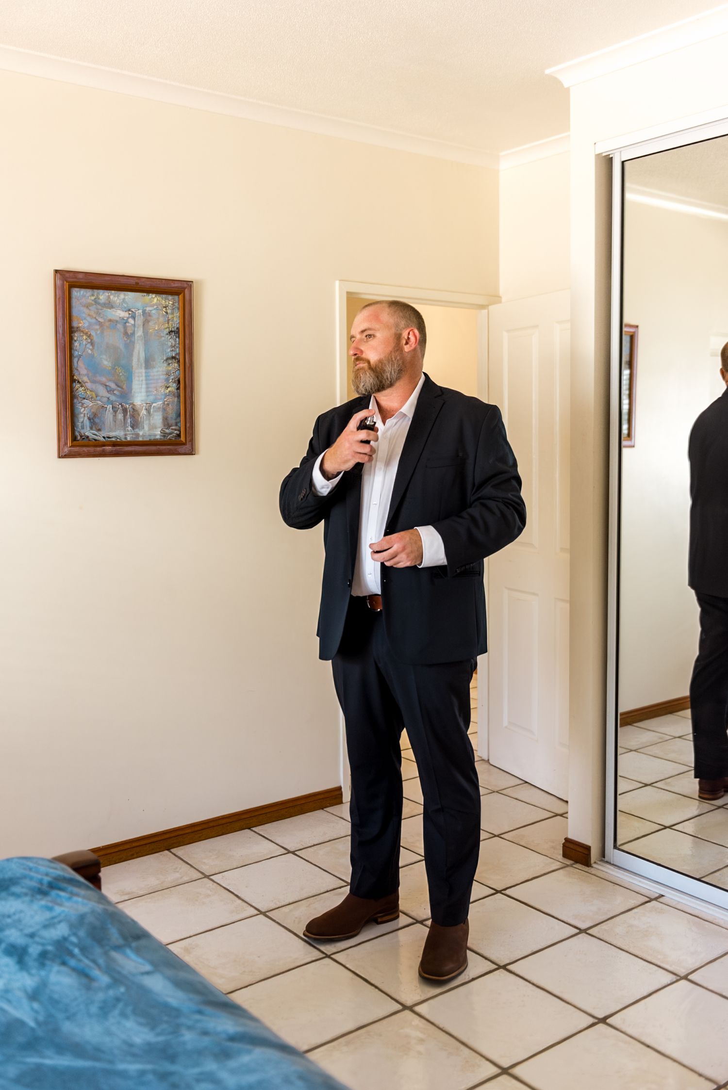Bearded groom in navy suit adjusting tie while standing in bedroom with mirror.