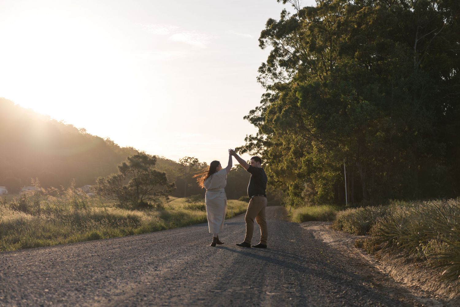 Couple dancing joyfully on rural gravel road surrounded by trees at sunset.