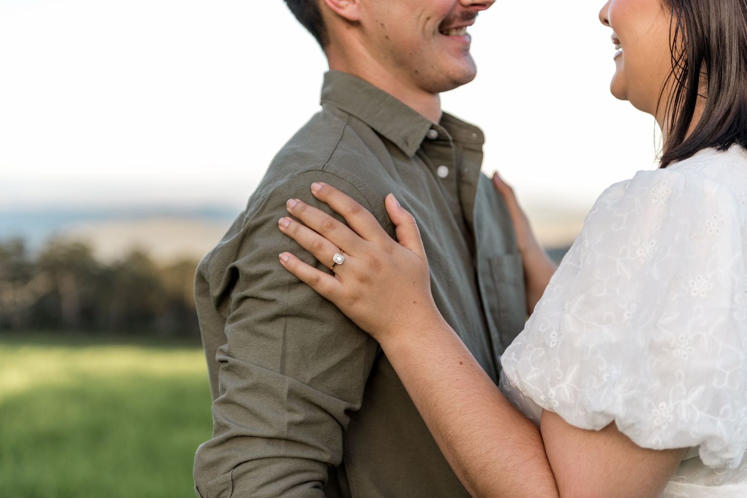 Close-up of woman's engagement ring as couple embraces in a green field.