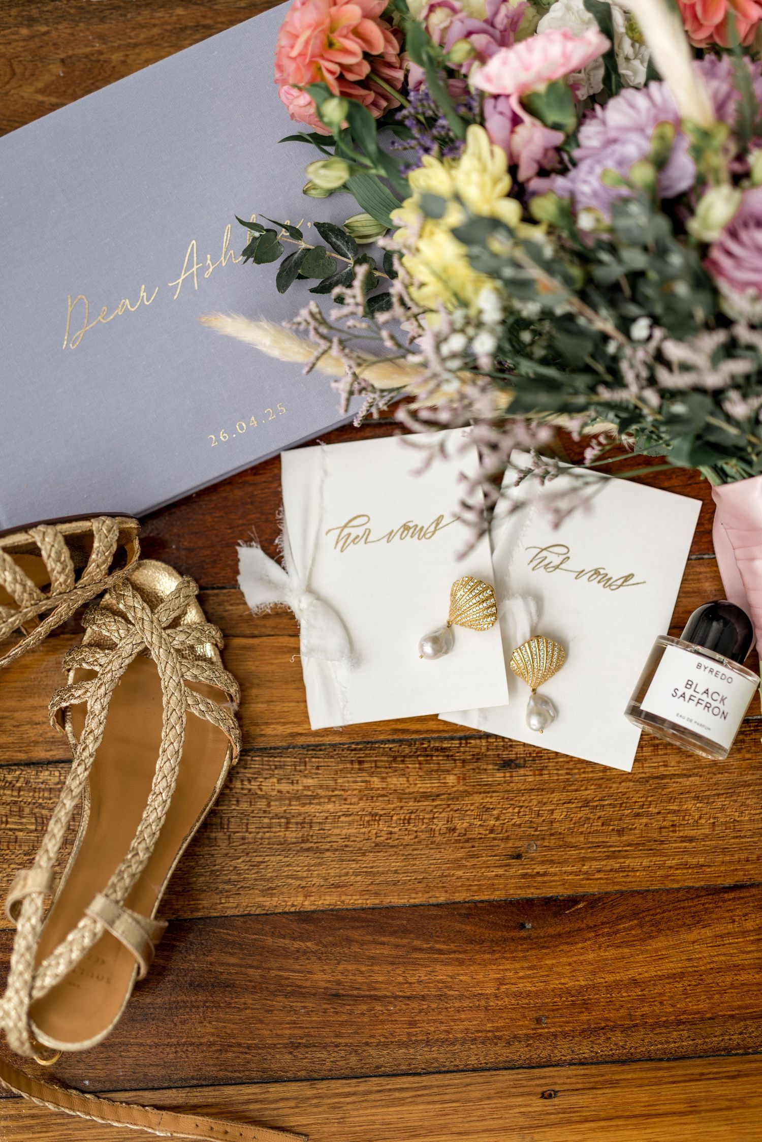 Gold shell earrings, handwritten vow cards, bridal shoes, and a floral bouquet arranged on a wooden table.