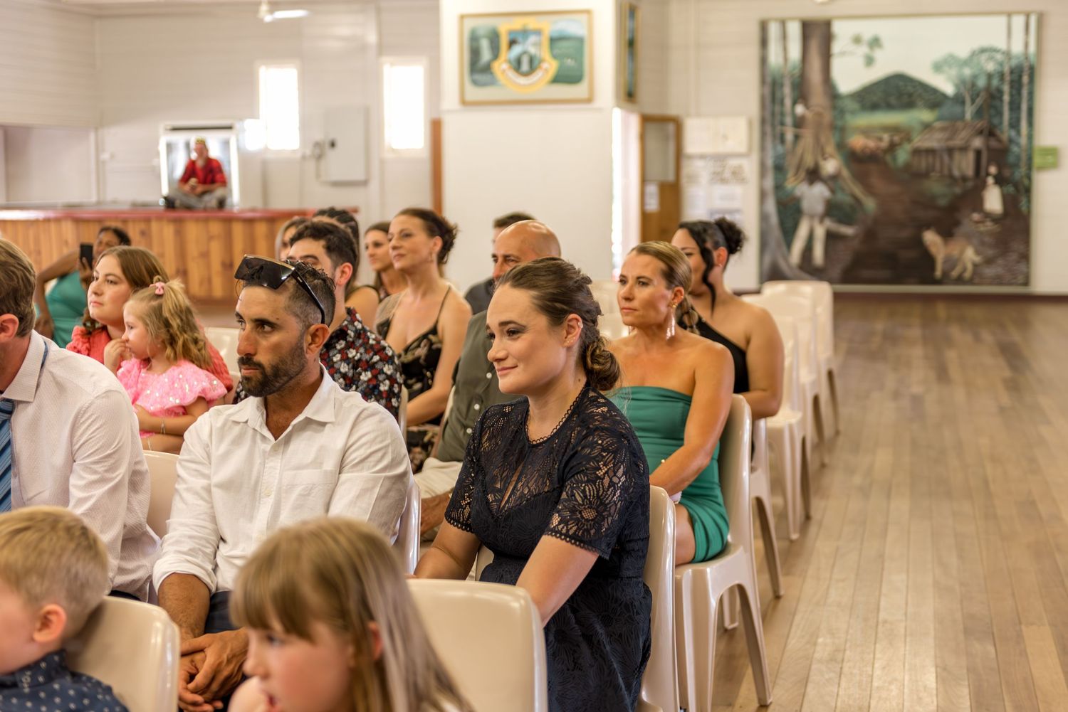 Wedding guests seated in white chairs watch ceremony inside rustic hall with colorful murals on the walls.