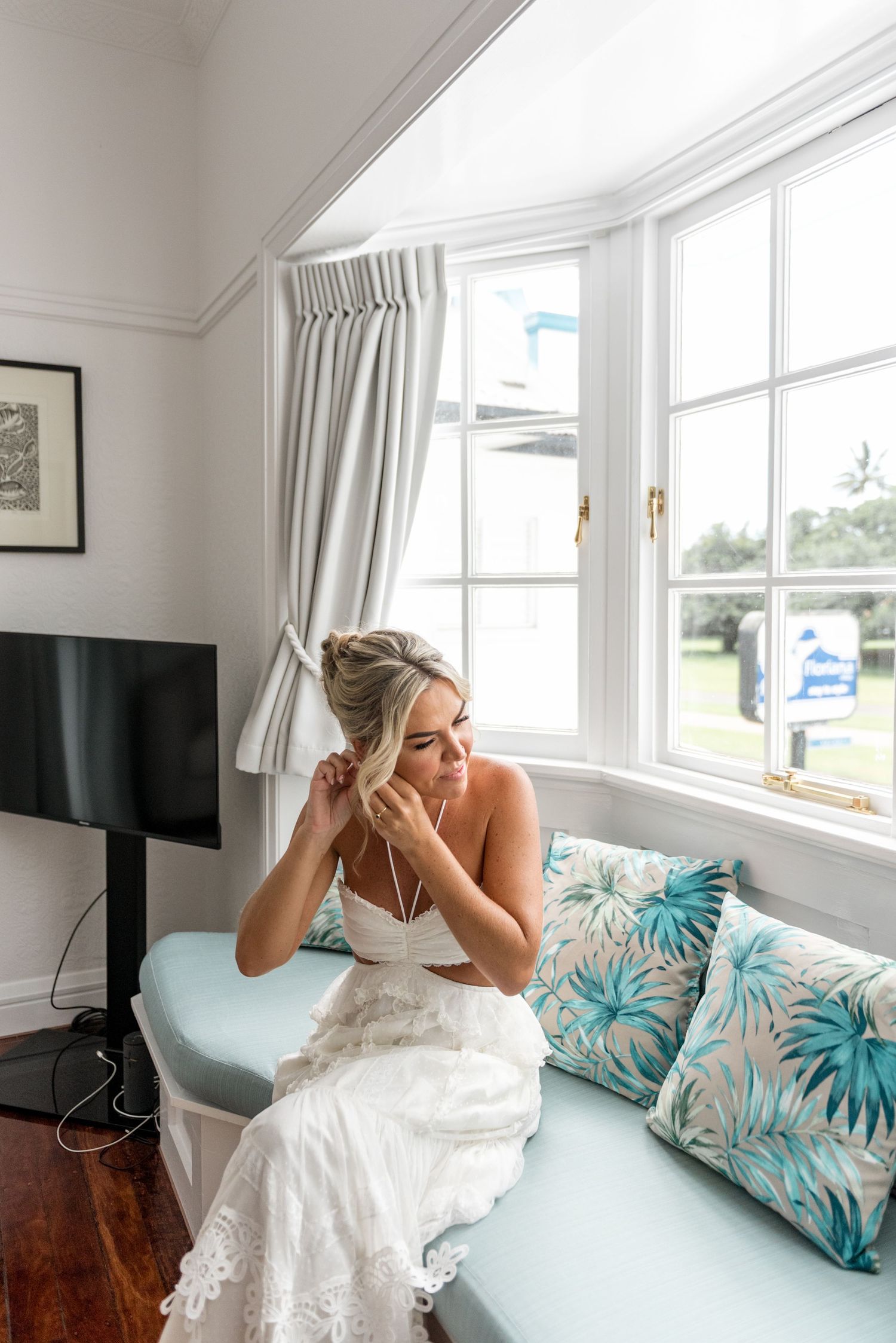 Elegant bride in white lace gown sitting by window adjusting earrings.