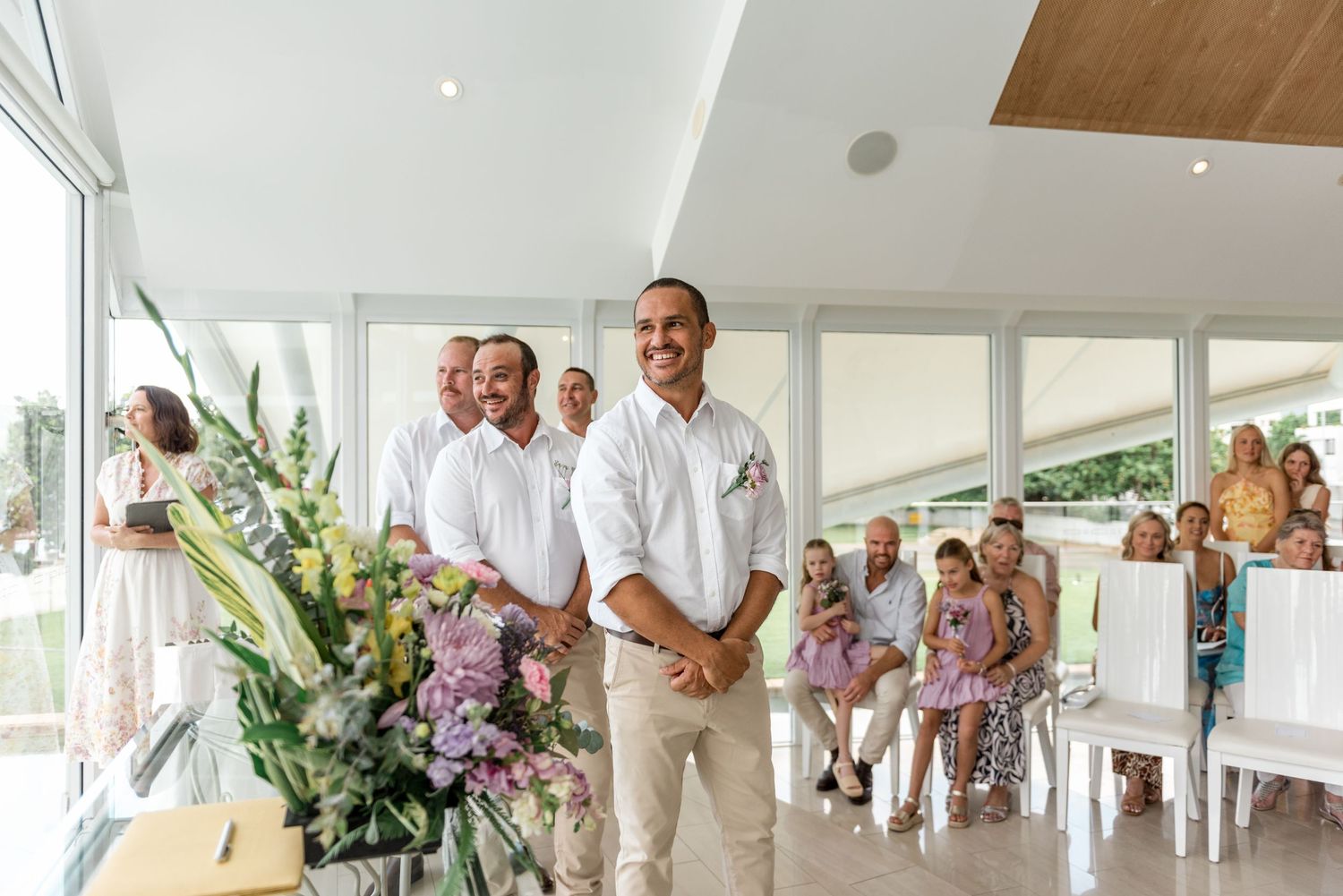 Groom standing at altar in white shirt smiling as guests watch the ceremony.