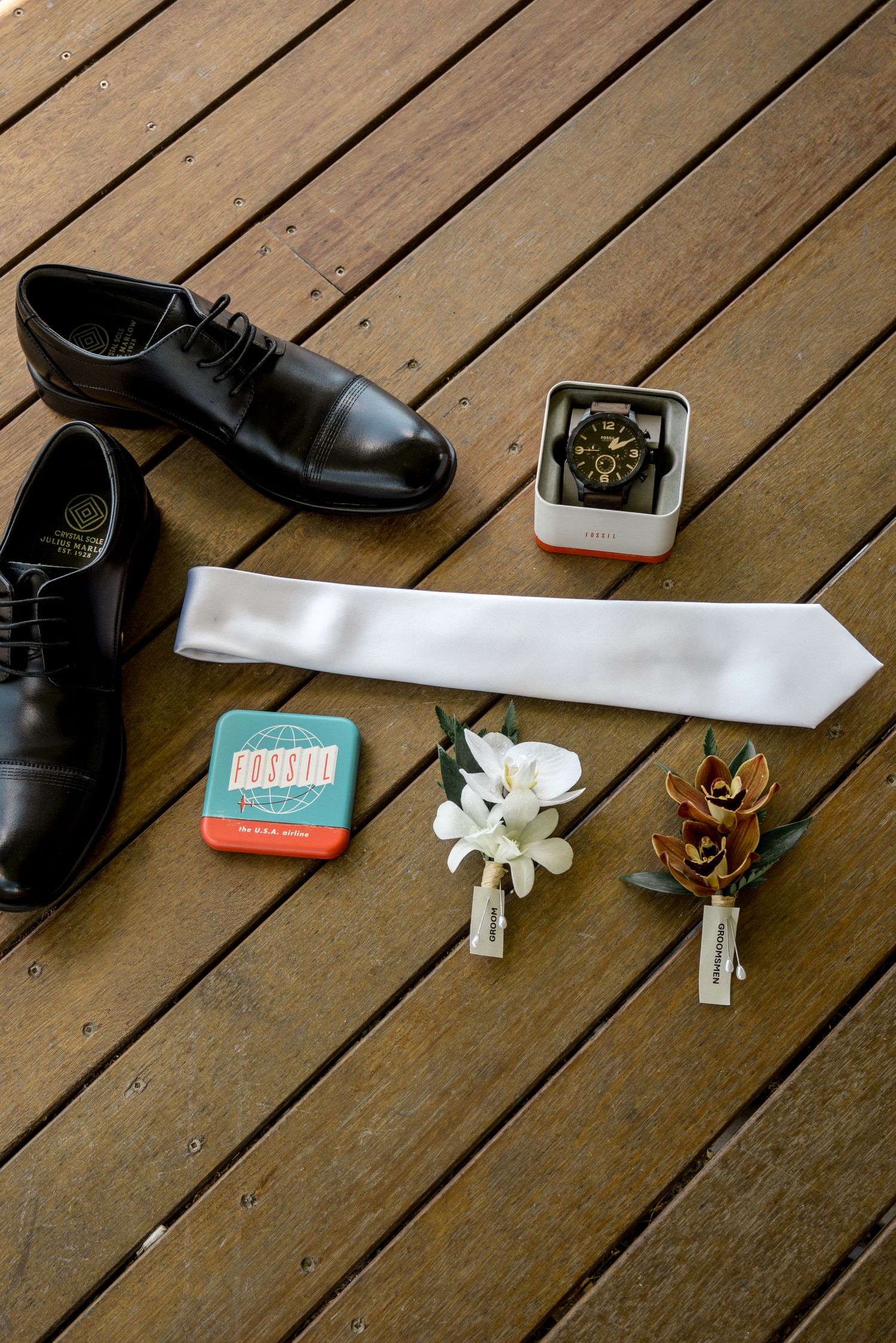 Grooms wedding flat lay featuring black dress shoes, white tie, watch, boutonniere, and accessories.