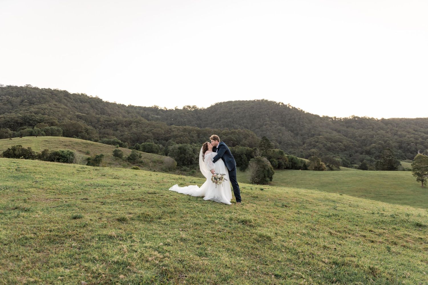 Groom dips bride in a romantic pose on a green hillside with beautiful rolling countryside in the background.