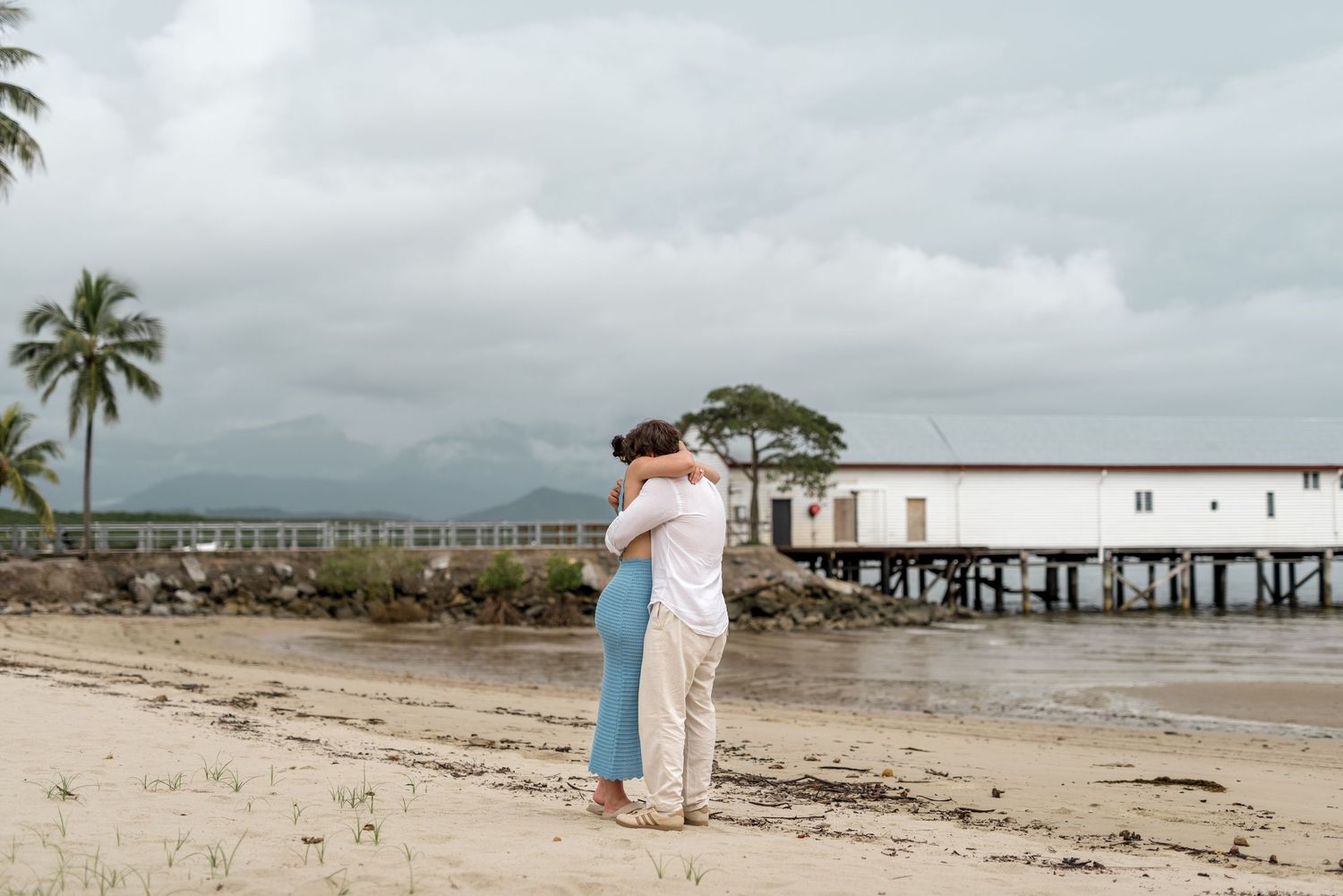 Couple hugging on sandy beach after proposal with scenic tropical waterfront pier backdrop.