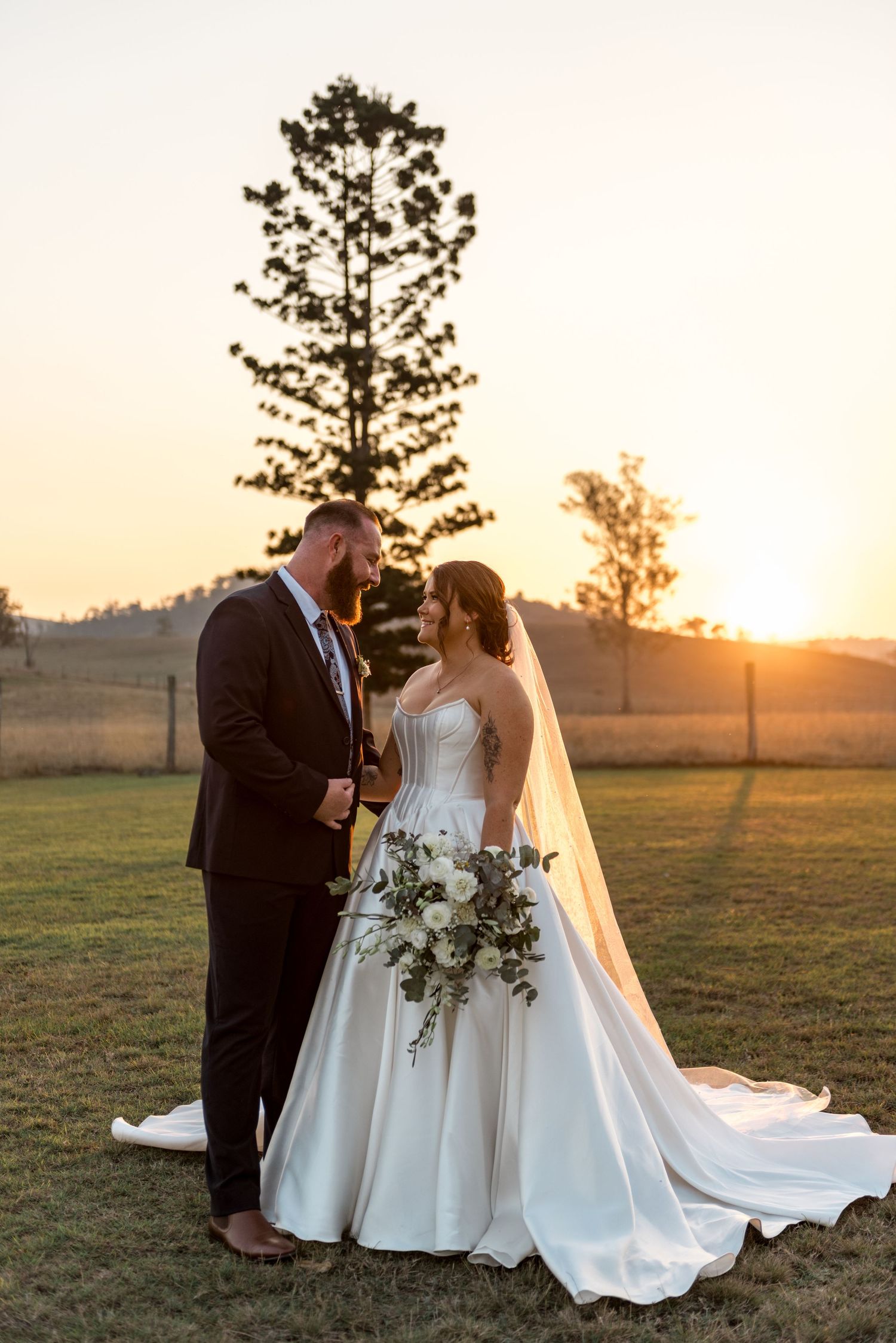 Bride and groom gazing at each other at sunset beside tall pine tree in open field.