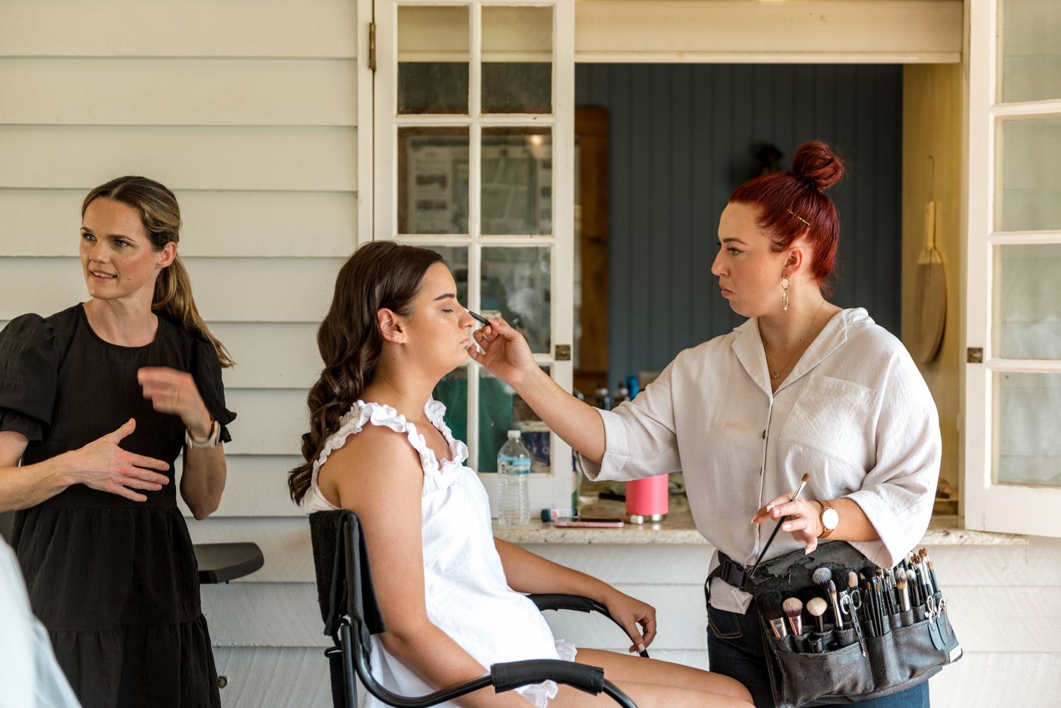 Bride getting makeup applied by red-haired artist on porch while bridesmaid looks on.