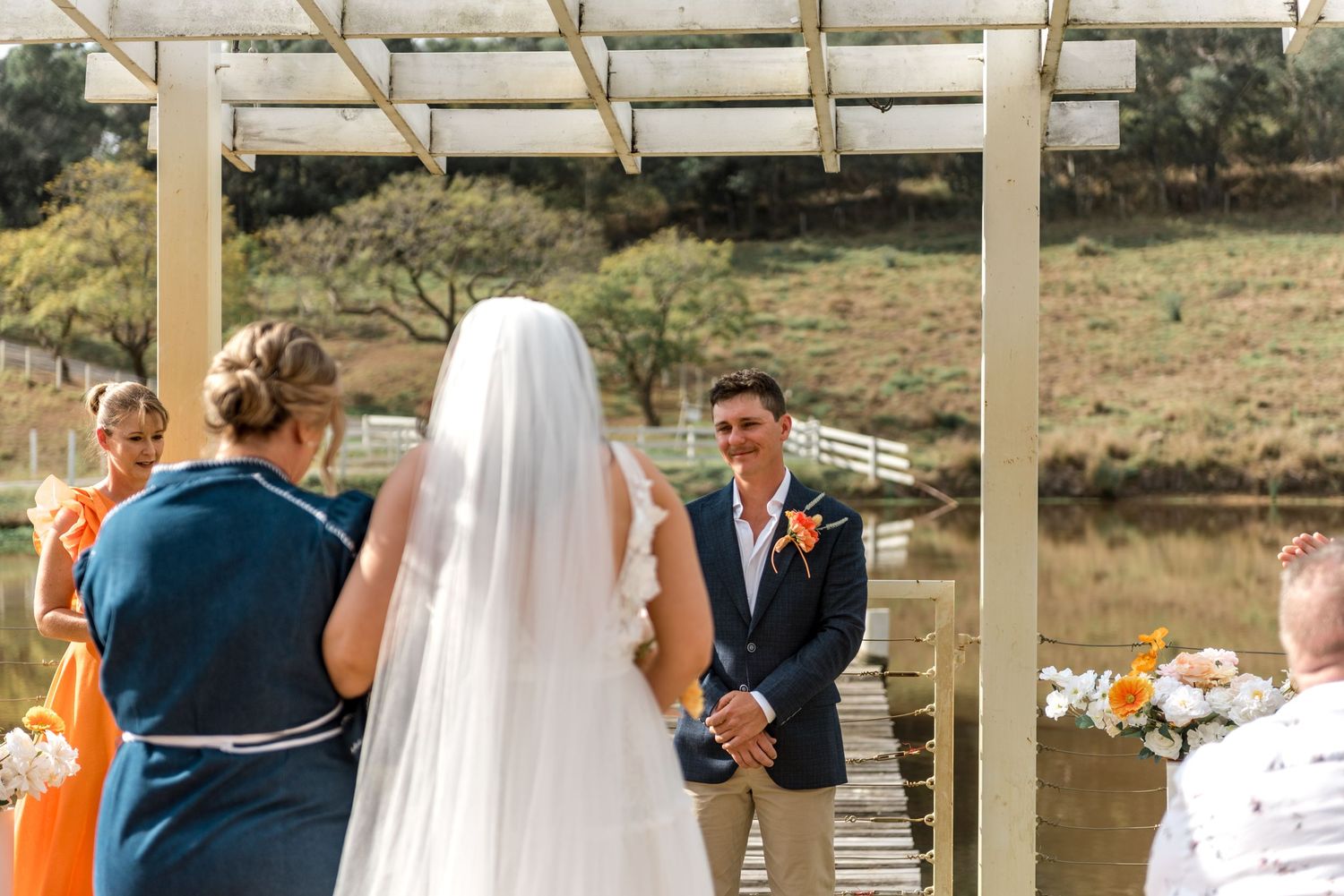 Bride in white gown and veil approaching smiling groom at outdoor wedding altar with pond backdrop.