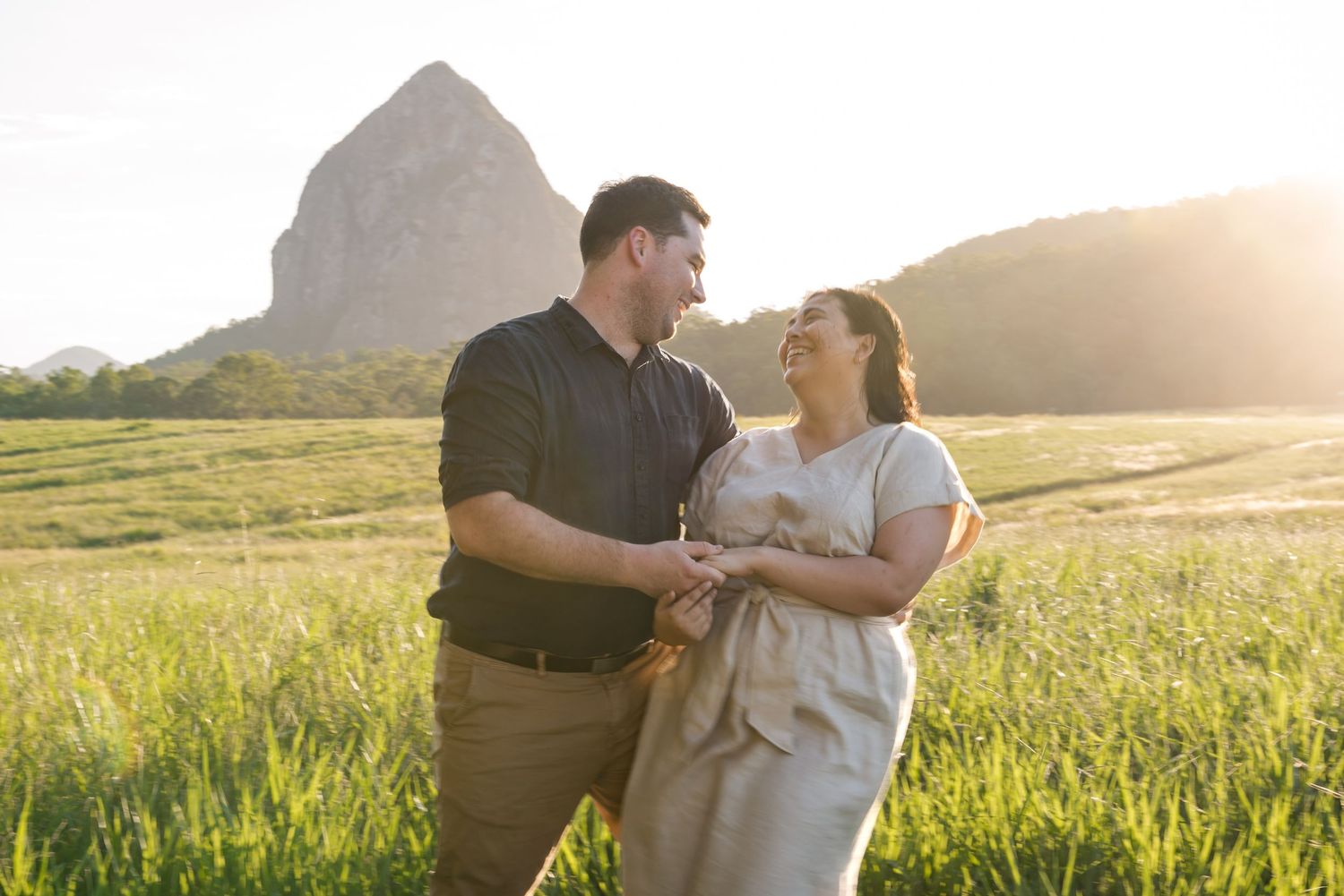 Couple holding hands laughing in green field with rocky mountain backdrop at golden hour.