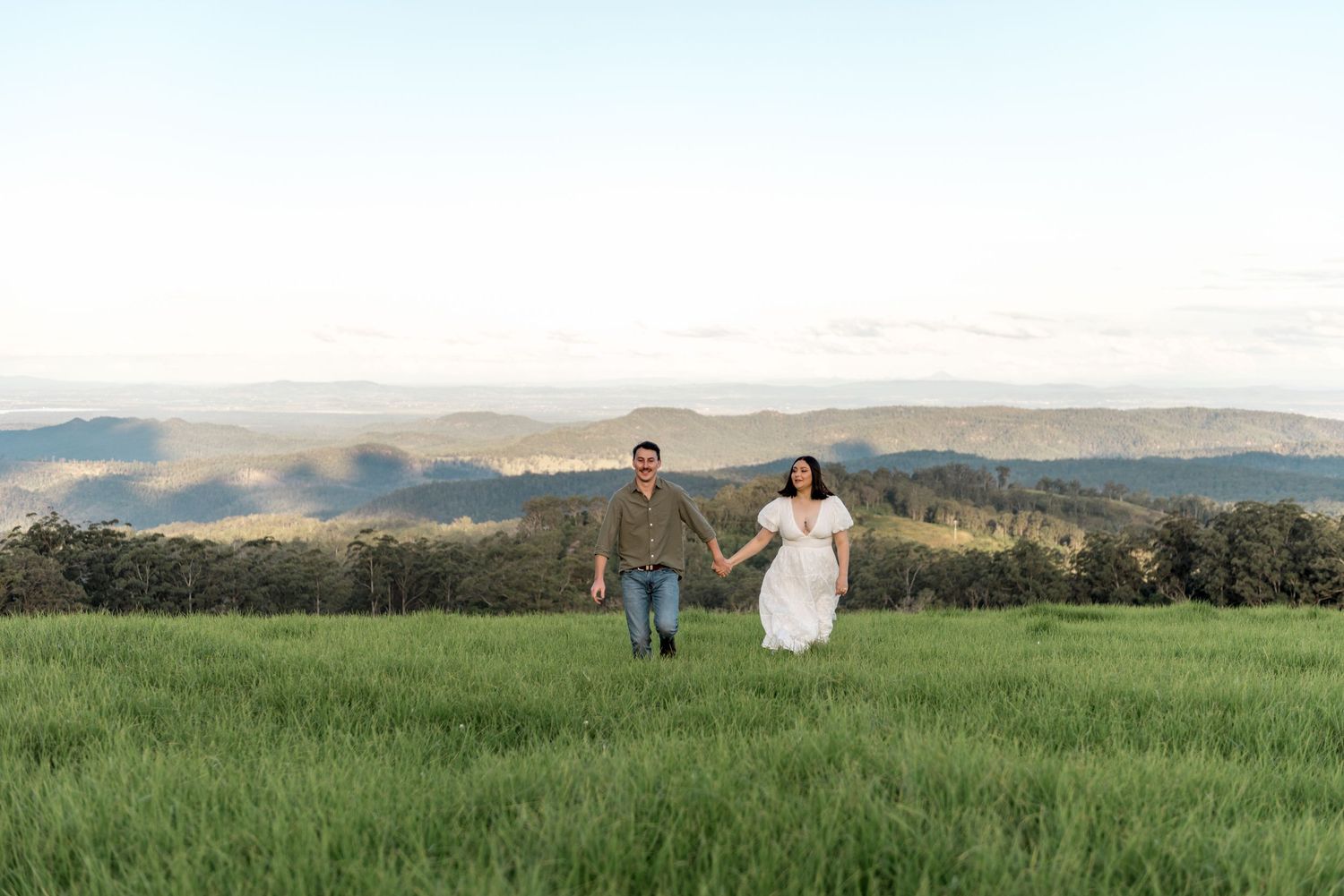 Happy couple holding hands running through a green meadow with mountain views.