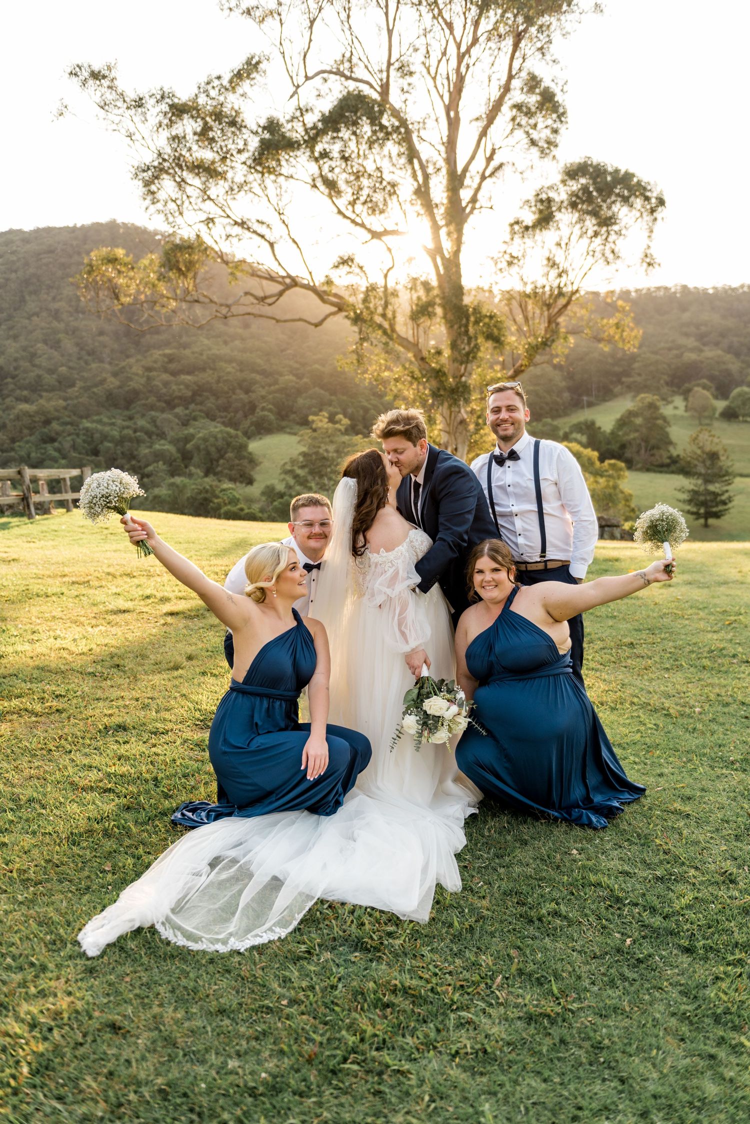 Wedding party celebrating on grass at sunset, bridesmaids in navy dresses raising bouquets.