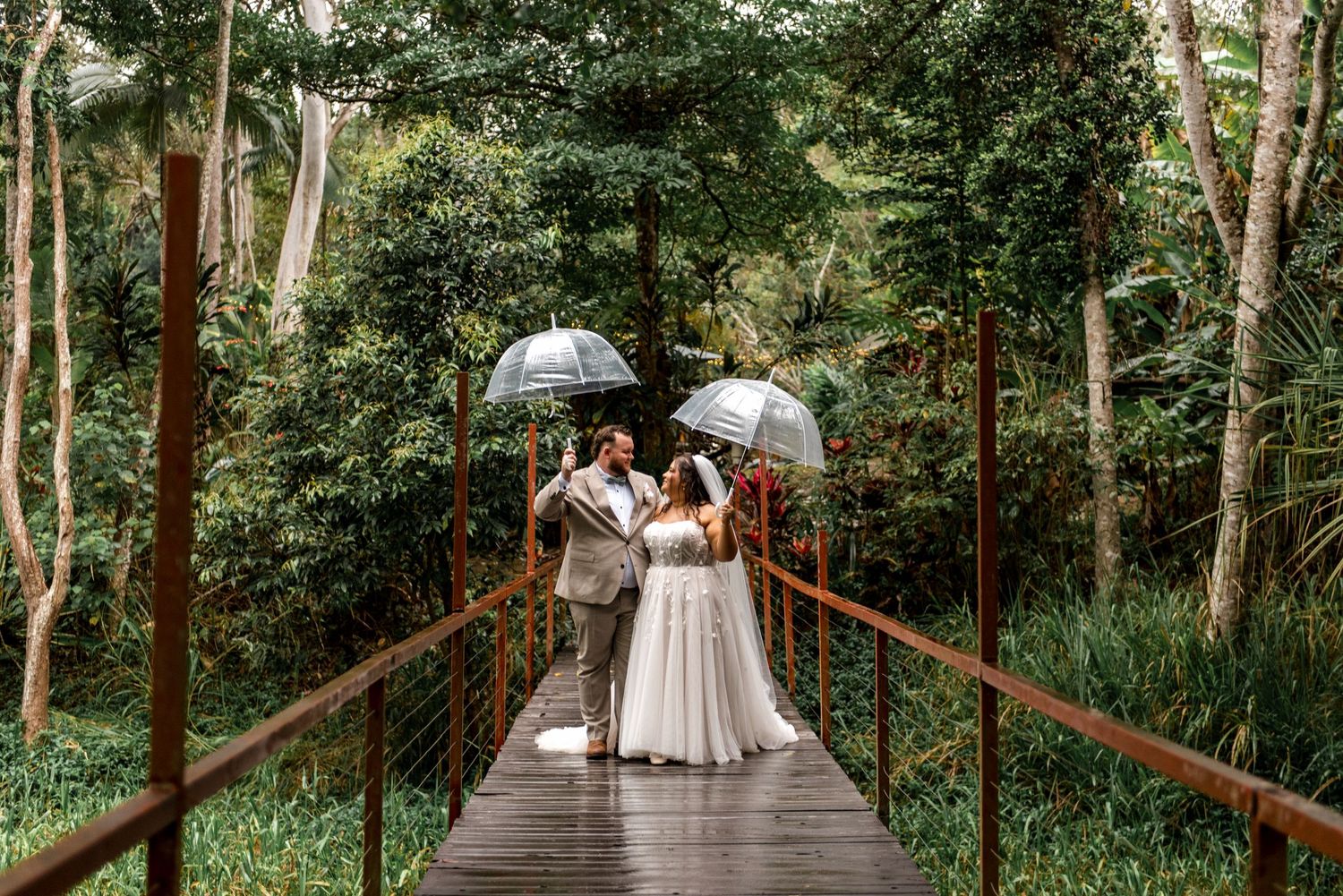 Bride and groom share an umbrella on a wooden bridge surrounded by dense tropical forest greenery.