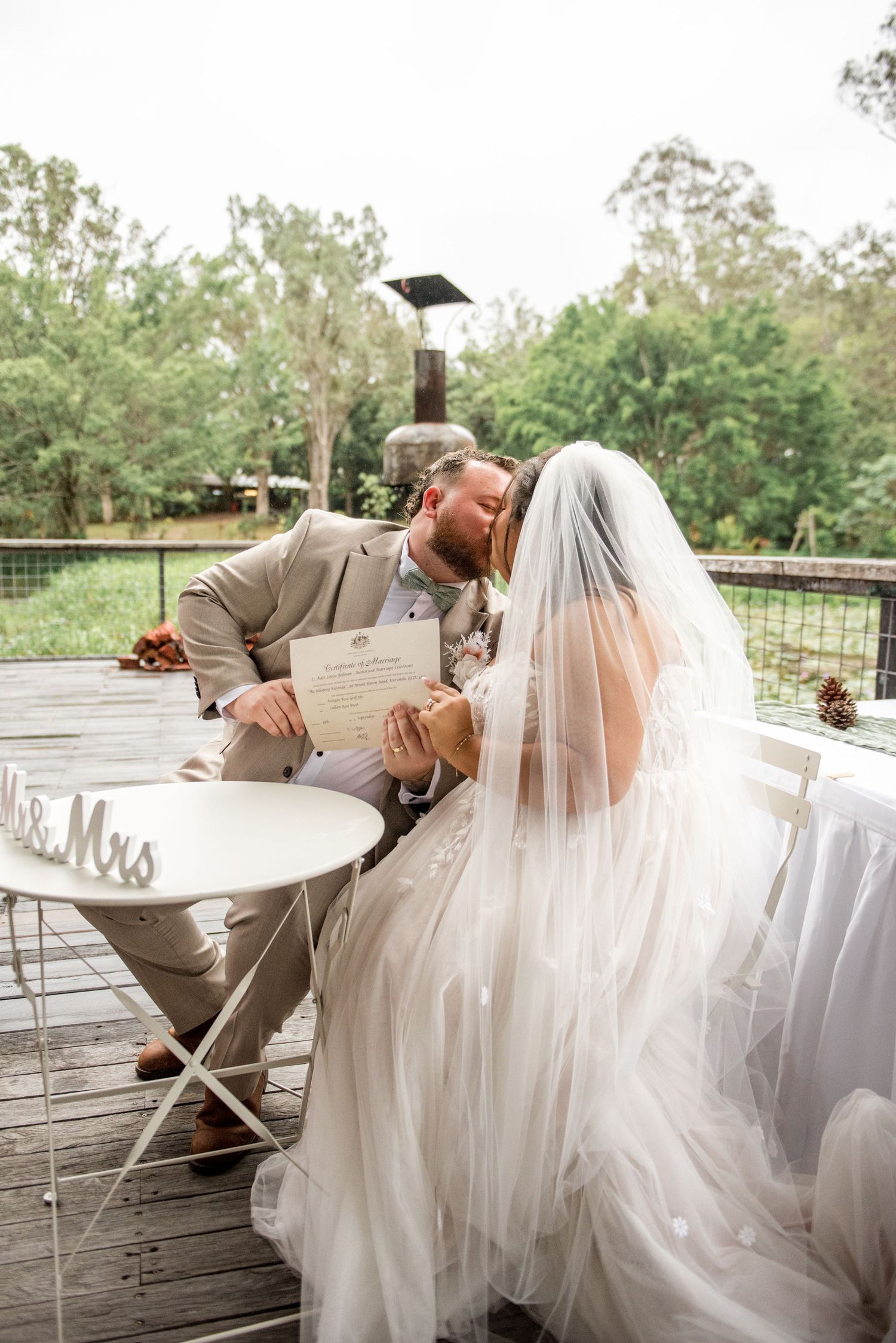 Bride and groom share a kiss while signing wedding certificate at a white table on a wooden deck.