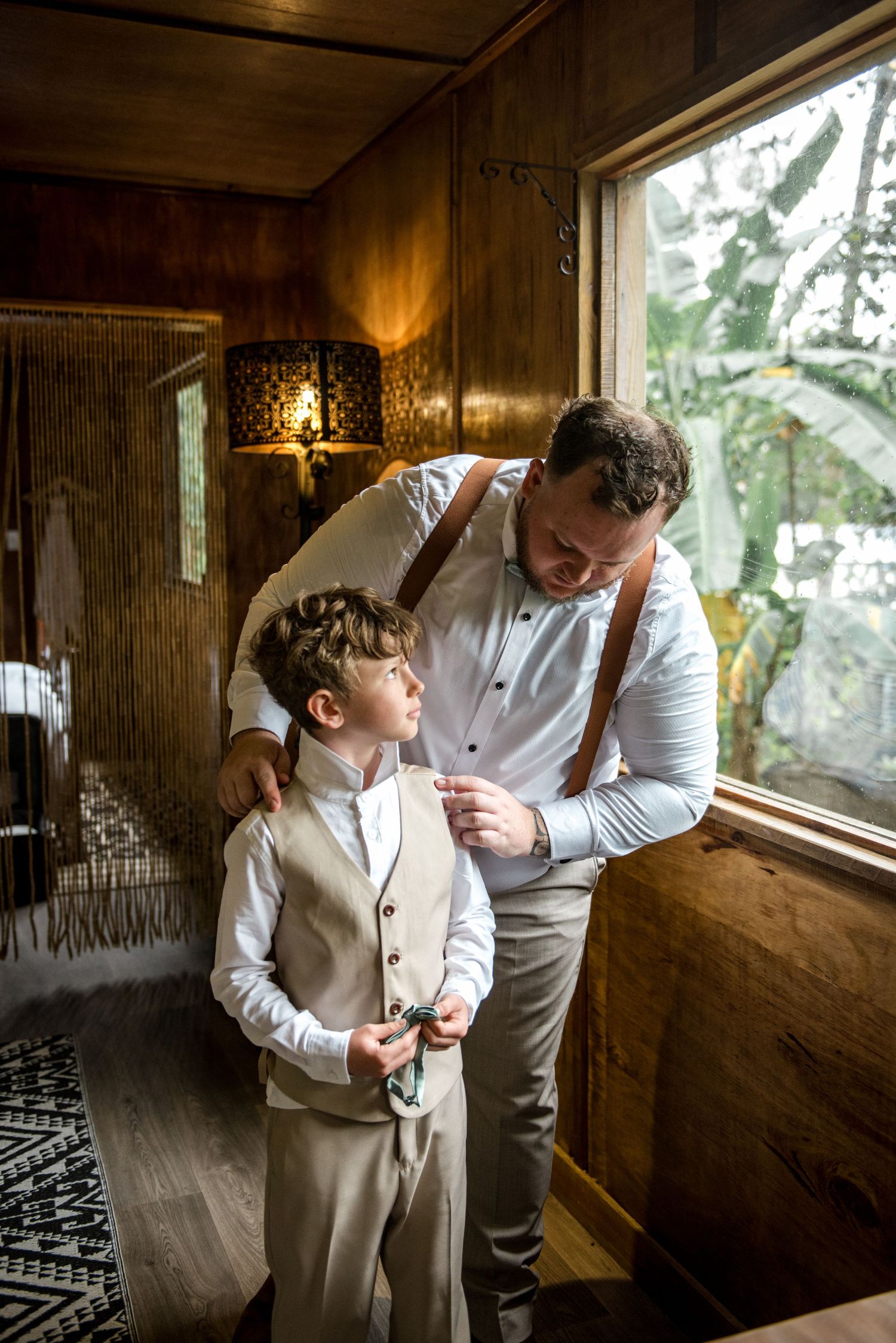 Groom in suspenders smiling down at young boy in beige suit while helping him get dressed.