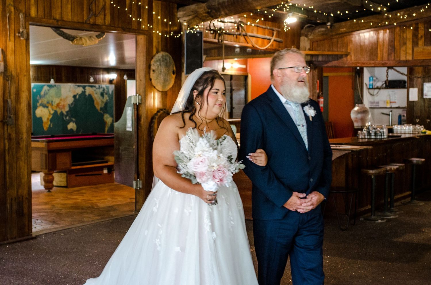 Bride in white gown walks down aisle with older man in navy suit at rustic indoor venue.