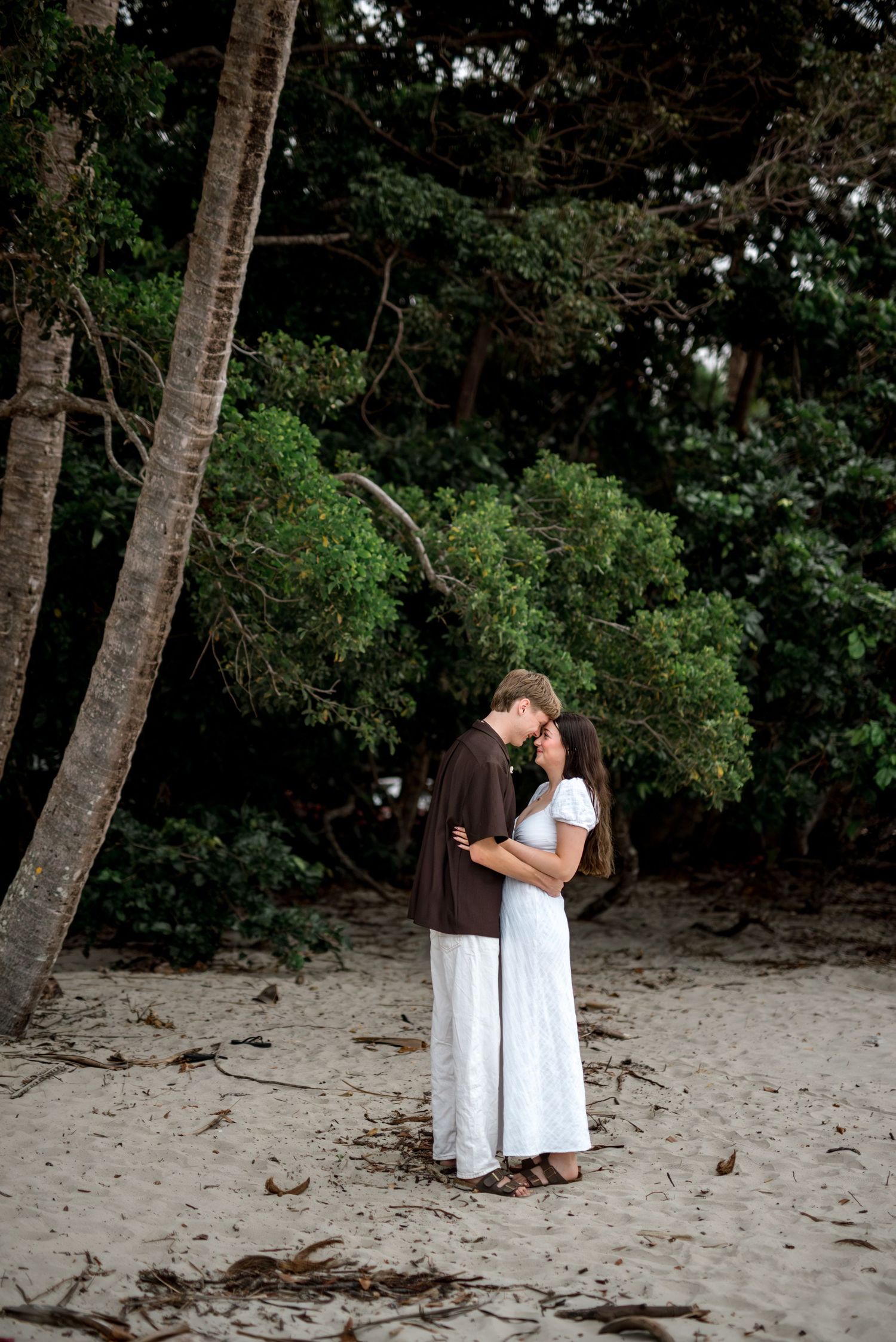 Romantic couple embracing and about to kiss on a sandy beach surrounded by tropical greenery.