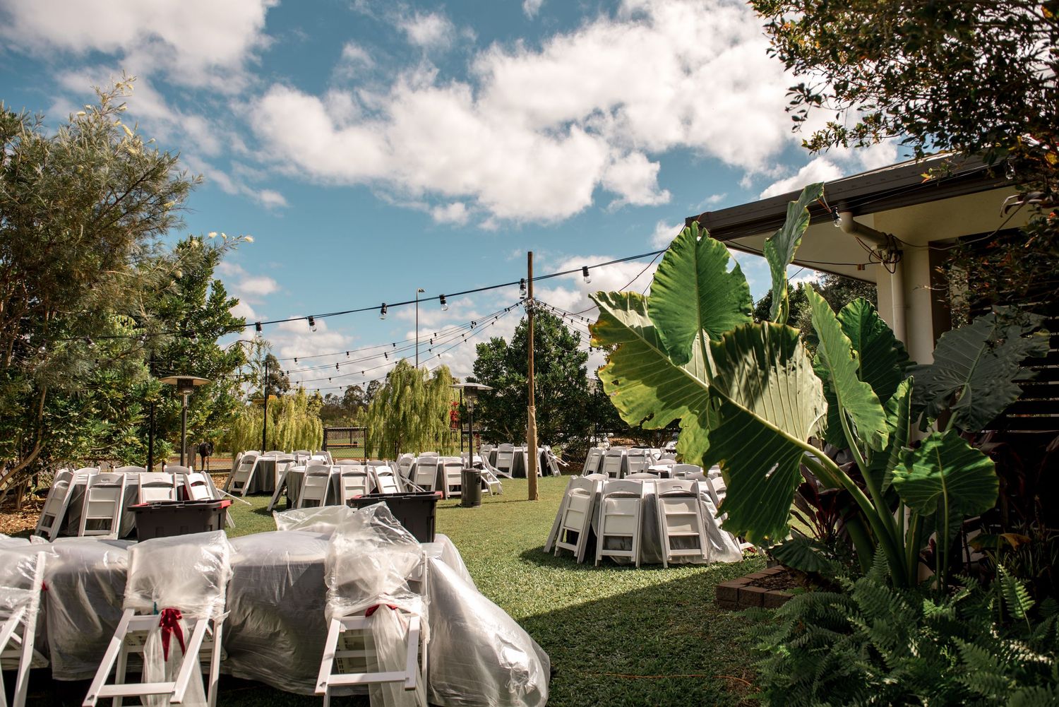 Outdoor wedding reception area with festoon lights, gray draped tables, white chairs, and tropical garden surroundings.