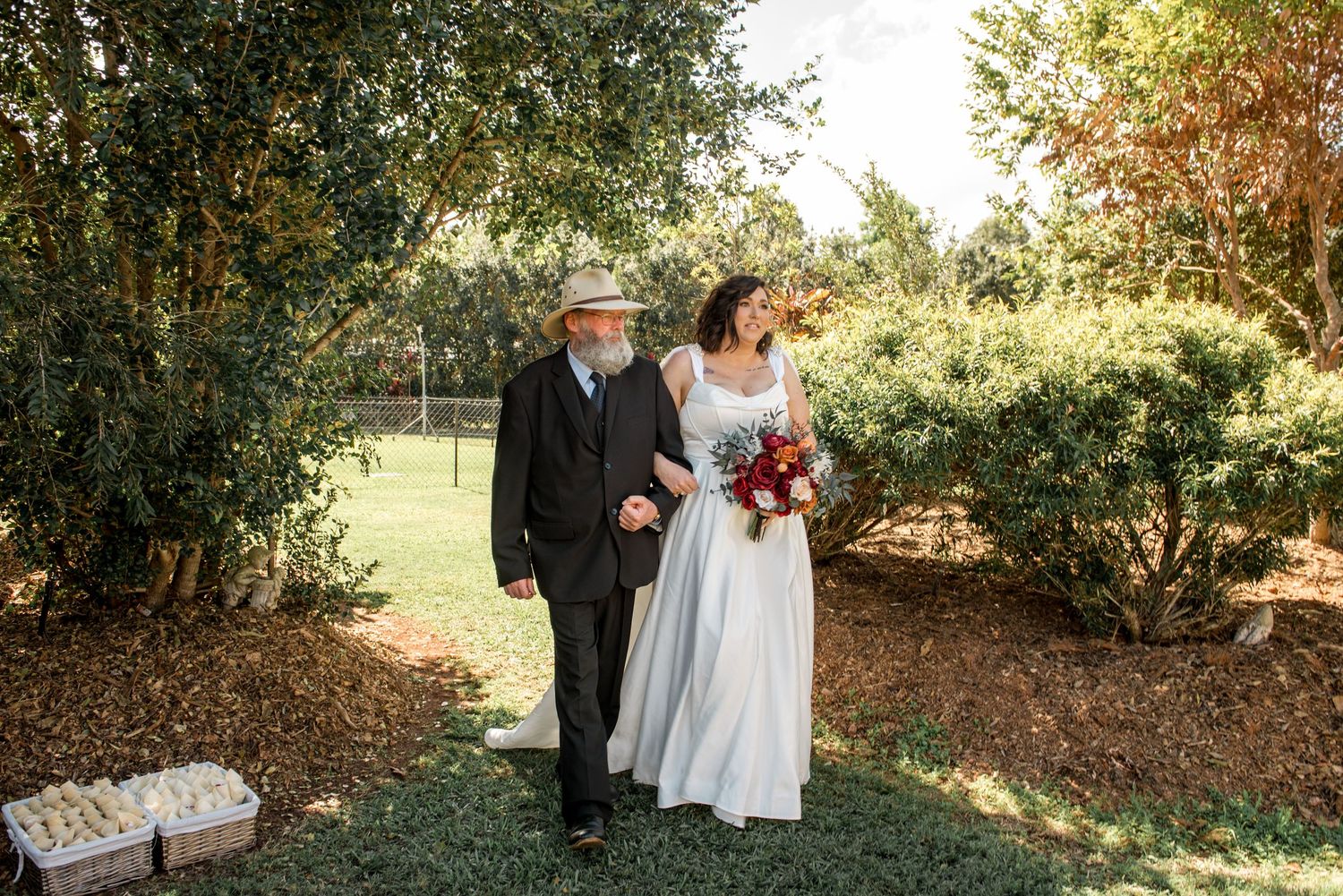 Bride in white gown and older man in hat stroll together through a lush green garden.