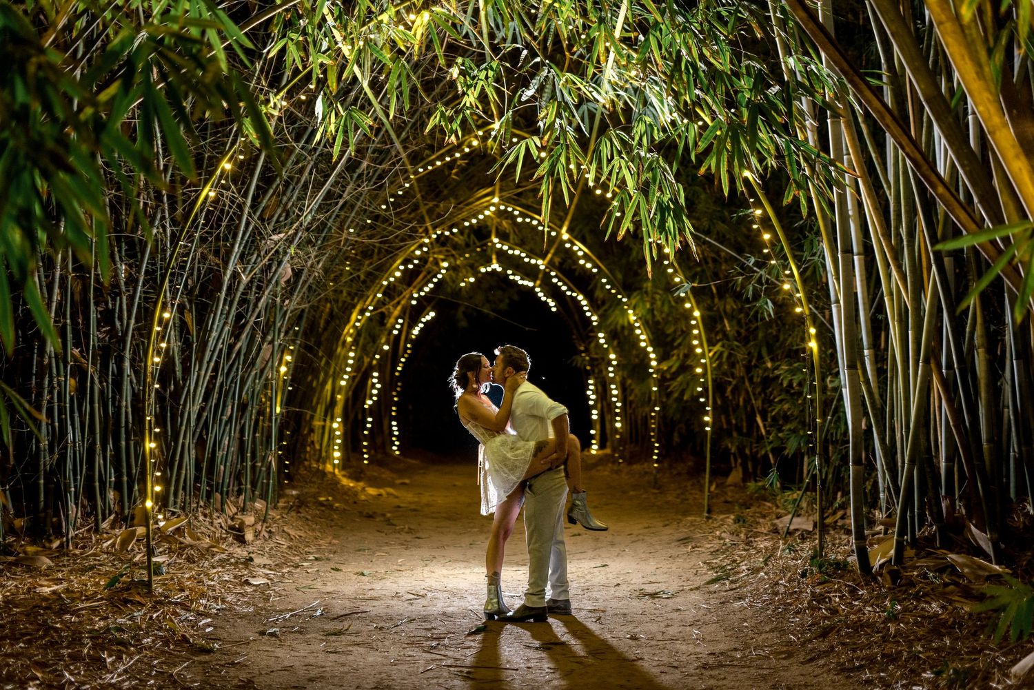 Couple sharing a romantic kiss in a bamboo tunnel adorned with warm fairy lights at night.