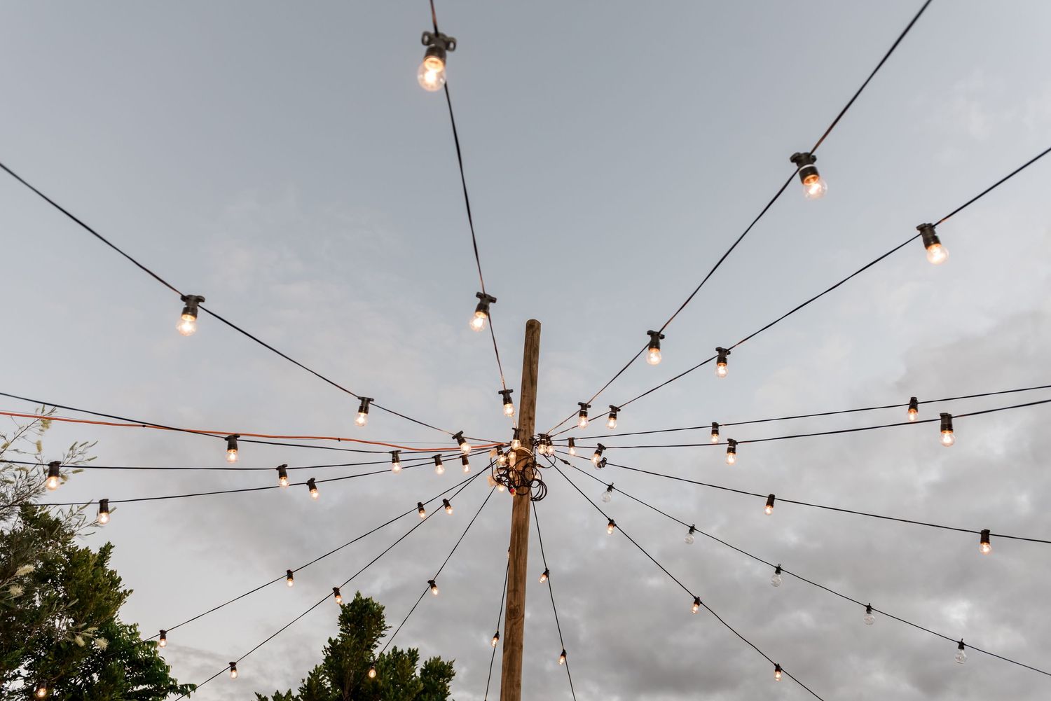 String lights radiating from central wooden pole against cloudy sky at outdoor wedding venue.