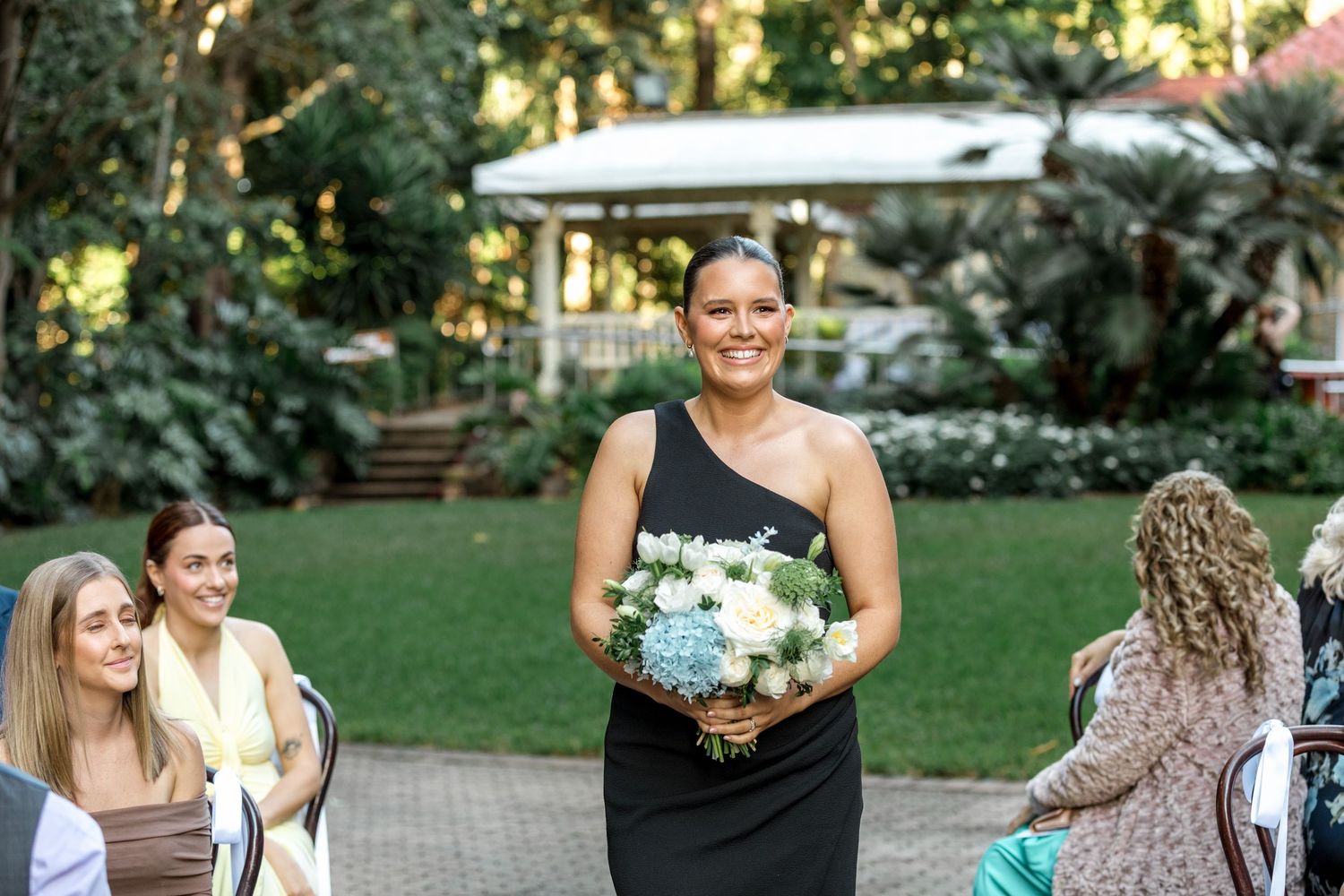 Bridesmaid in black one-shoulder dress carries white and blue bouquet down garden aisle.