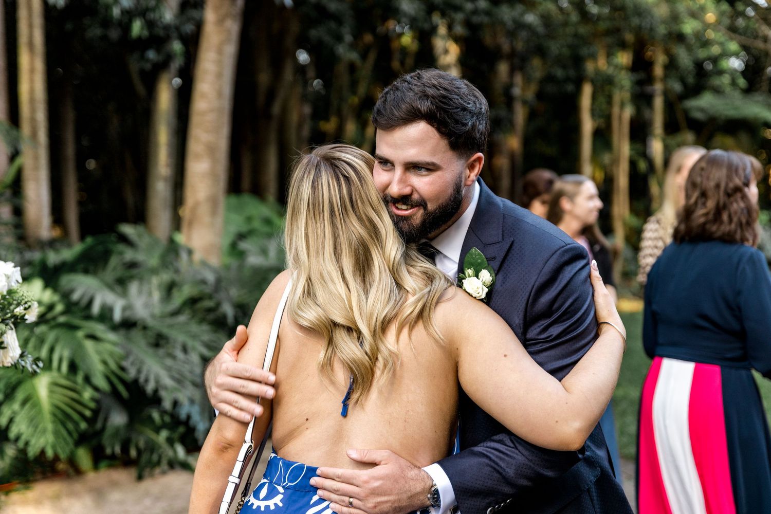Groom in navy suit warmly embraces a blonde female guest during outdoor garden wedding celebration.
