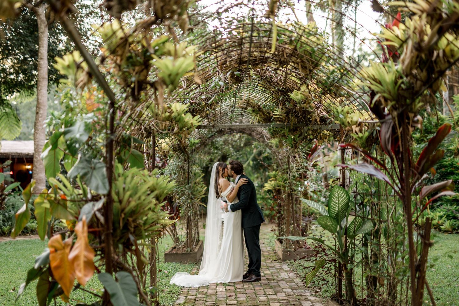 Bride and groom kiss under a stunning botanical archway in a tropical garden.