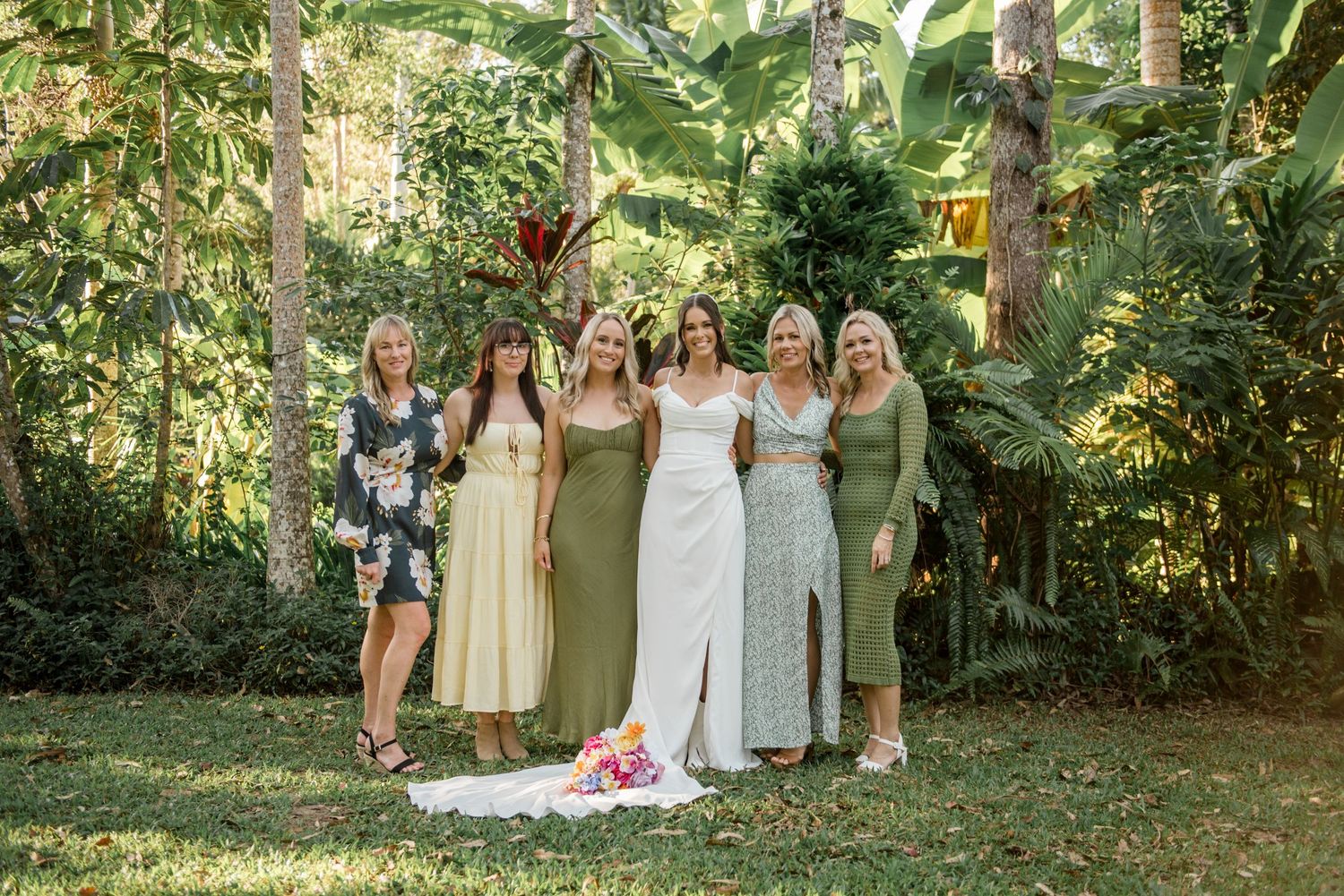 Bride in white gown poses with four bridesmaids in green and neutral dresses in tropical garden.