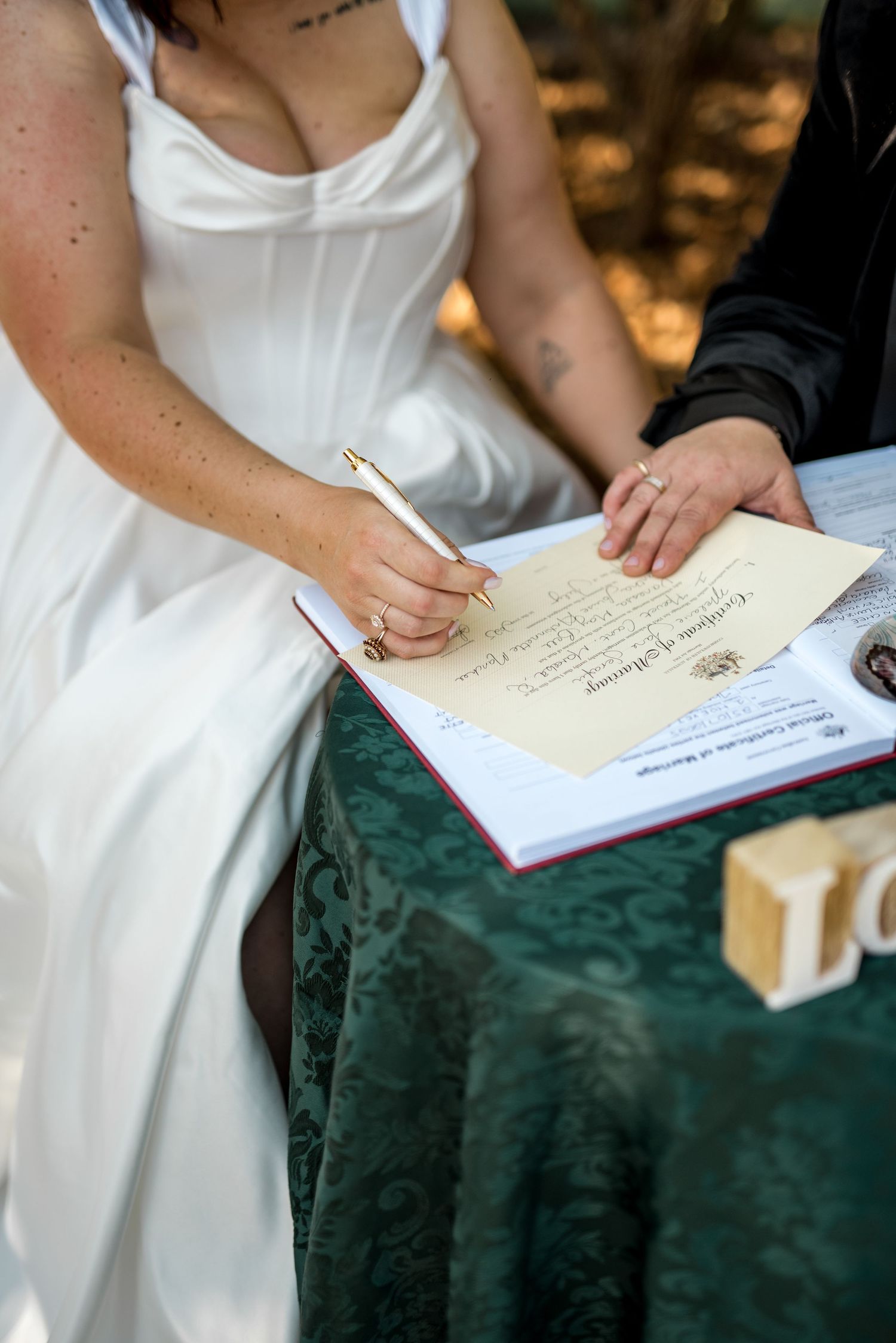 Bride in white gown signs marriage certificate at green velvet table during wedding ceremony.