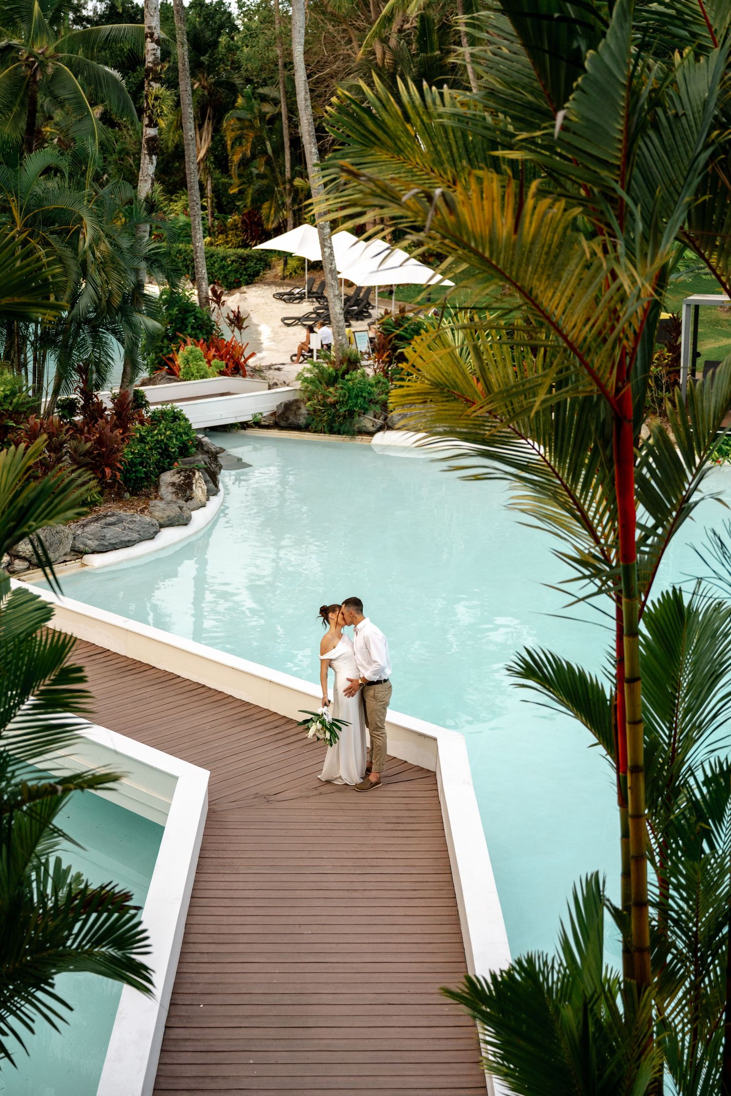 Aerial view of bride and groom kissing on a wooden deck beside a lagoon-style resort pool at Sheraton Grand Mirage Port Douglas.