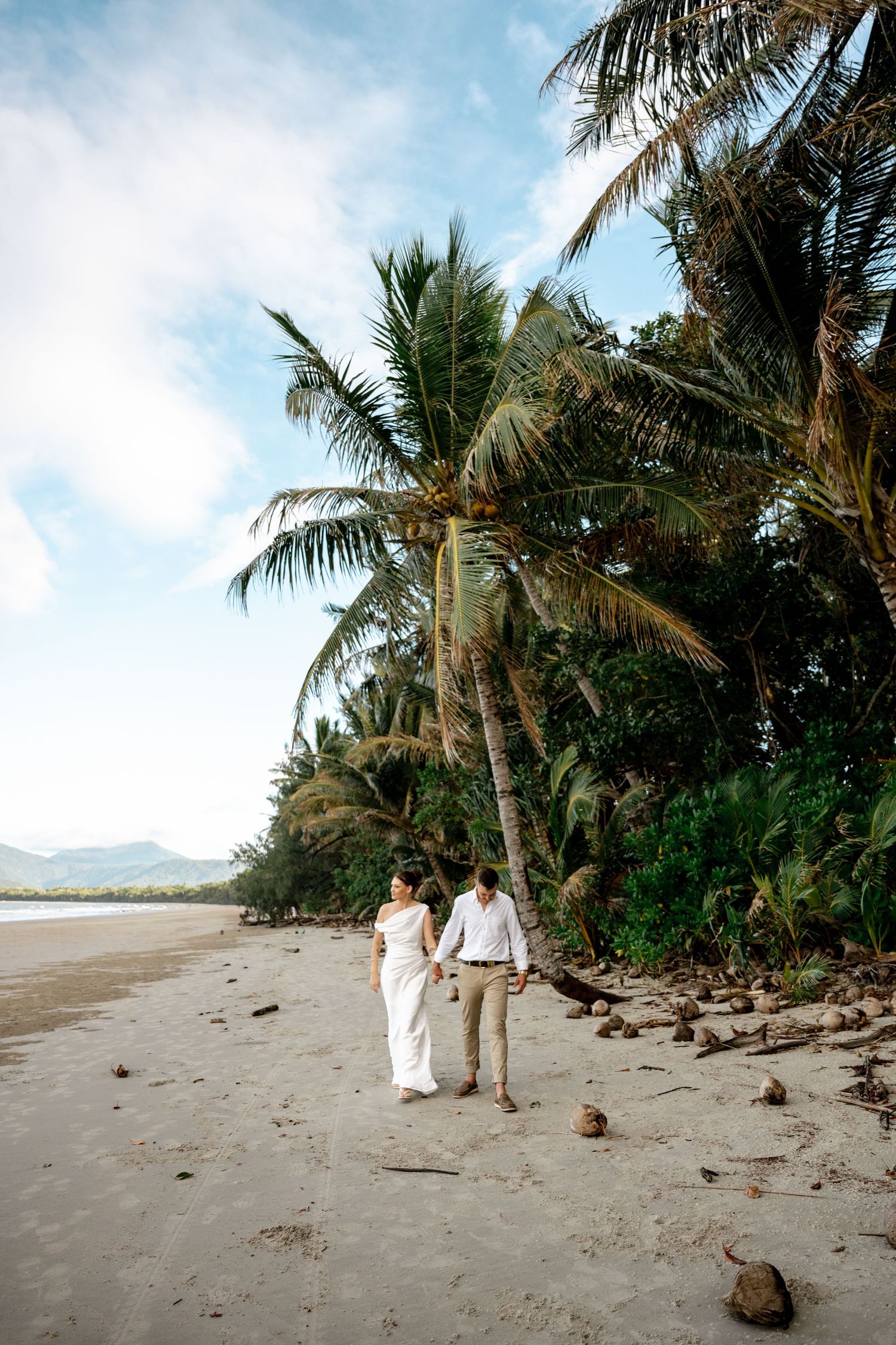 Bride and groom hold hands walking on sandy beach beneath towering tropical palm trees at Four Mile Beach, Port Douglas.