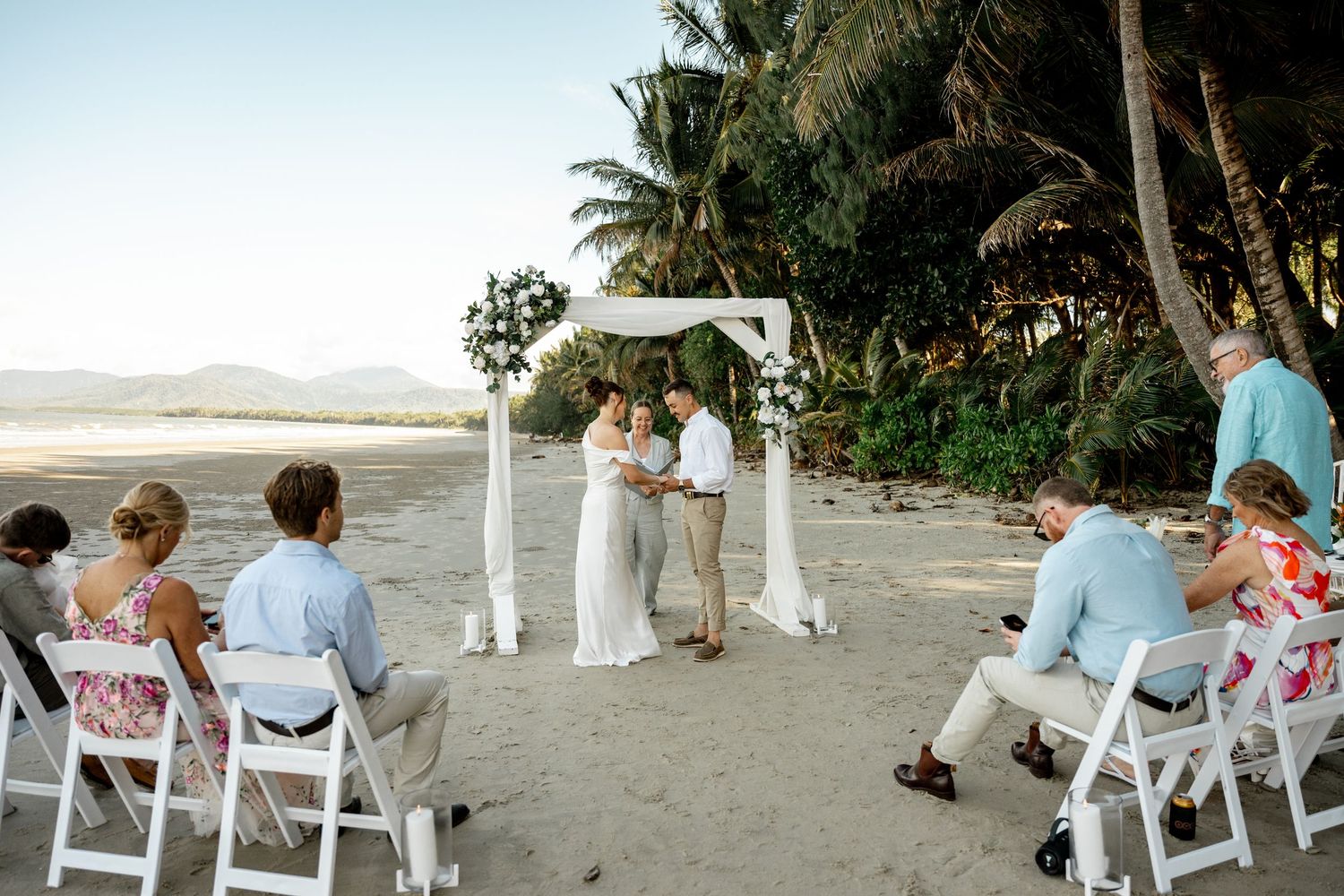 Small intimate beach wedding ceremony with guests seated on white chairs, couple at floral arch, tropical setting at Four Mile Beach.