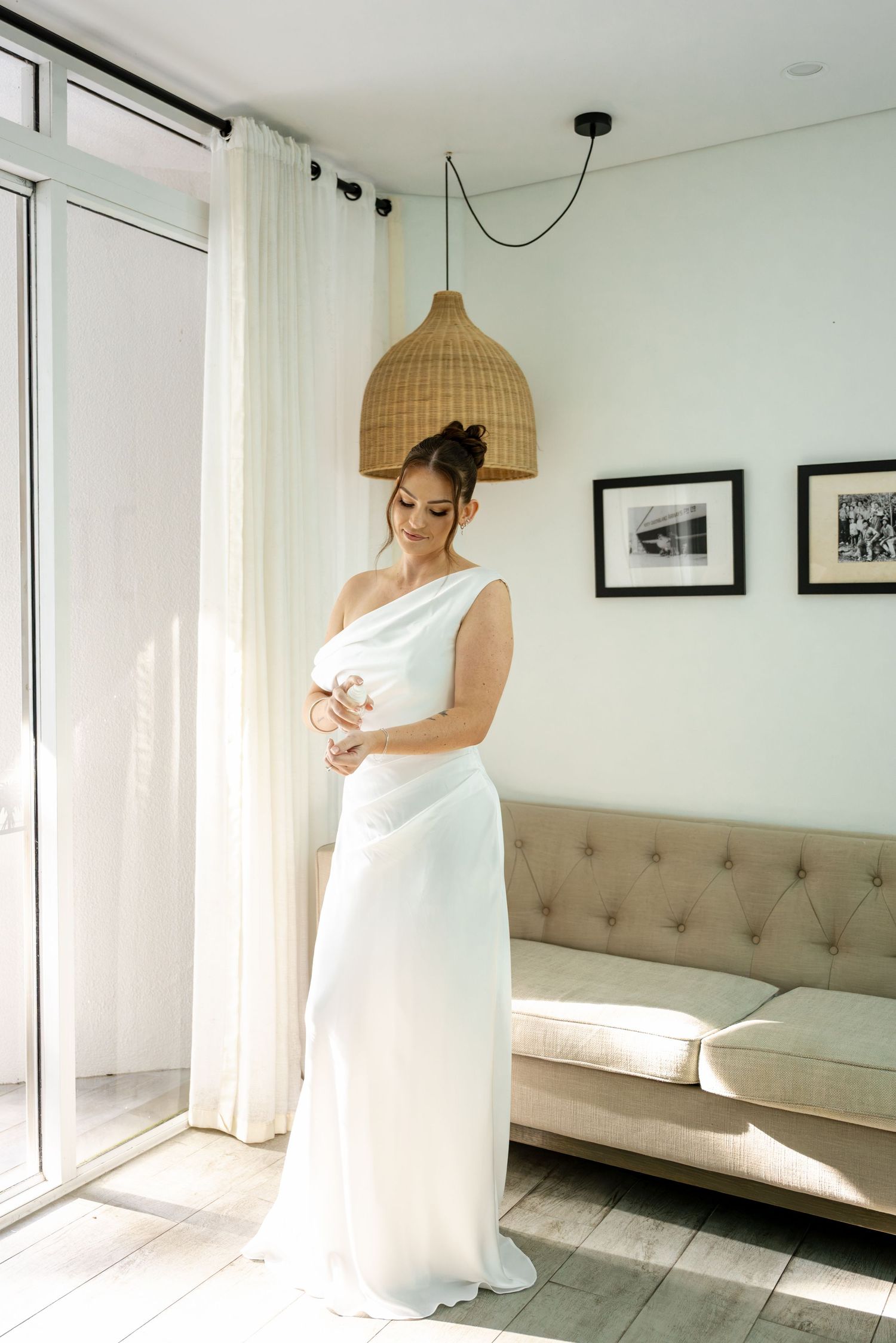 Elegant bride in one-shoulder white satin gown stands by window in sunlit room with rattan pendant lamp.
