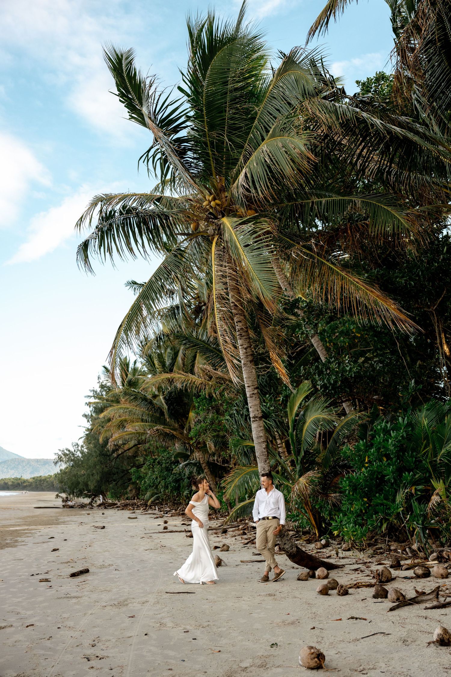 Wedding couple walks along sandy tropical beach lined with tall coconut palm trees at Four Mile Beach, Port Douglas.