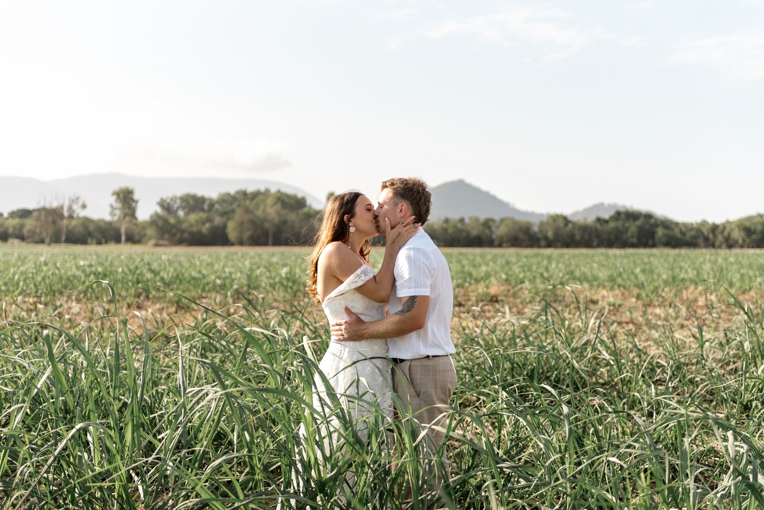 Romantic couple kissing in a green field with mountains and trees in the background.