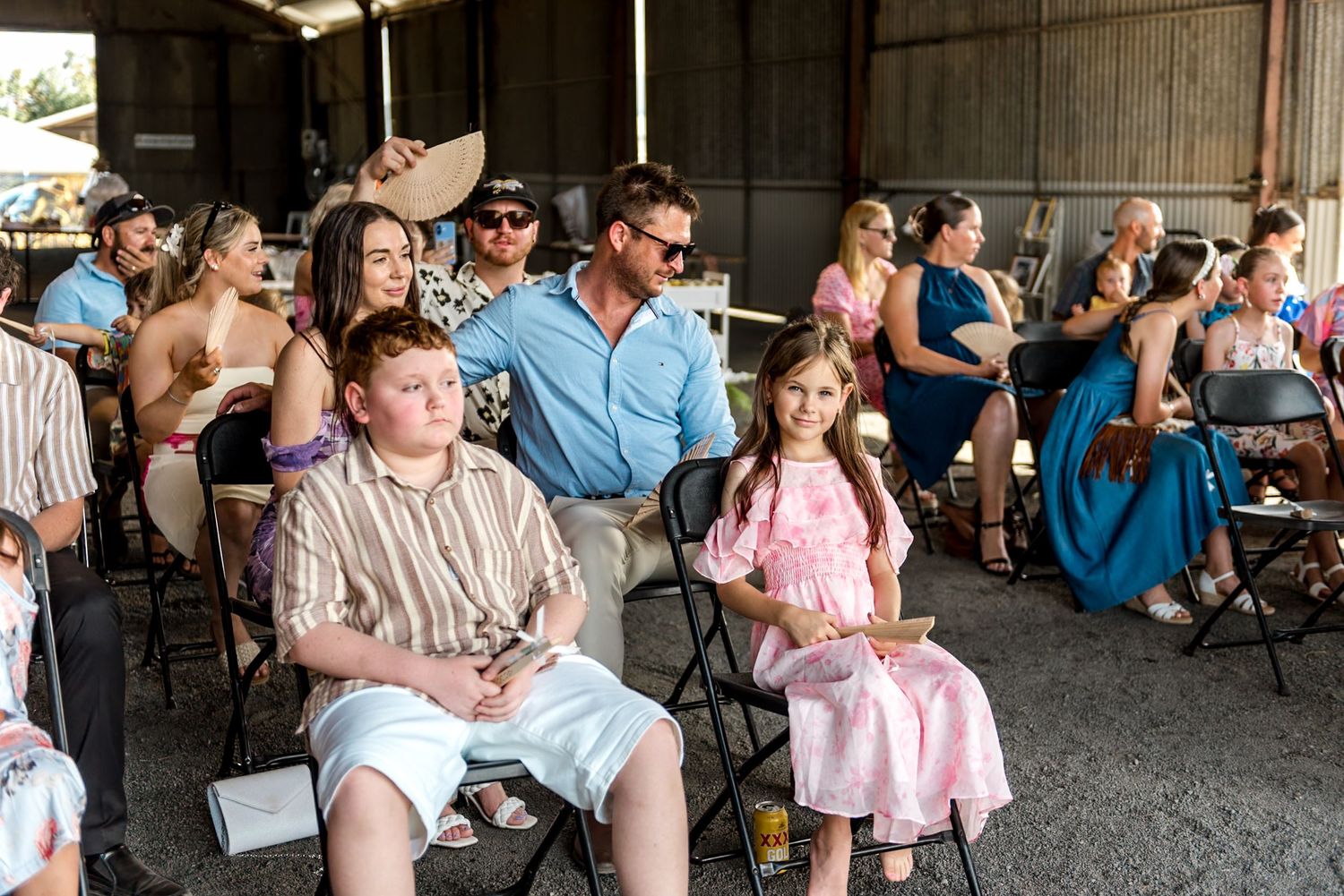 Wedding guests including children seated on folding chairs inside a rustic barn ceremony venue.