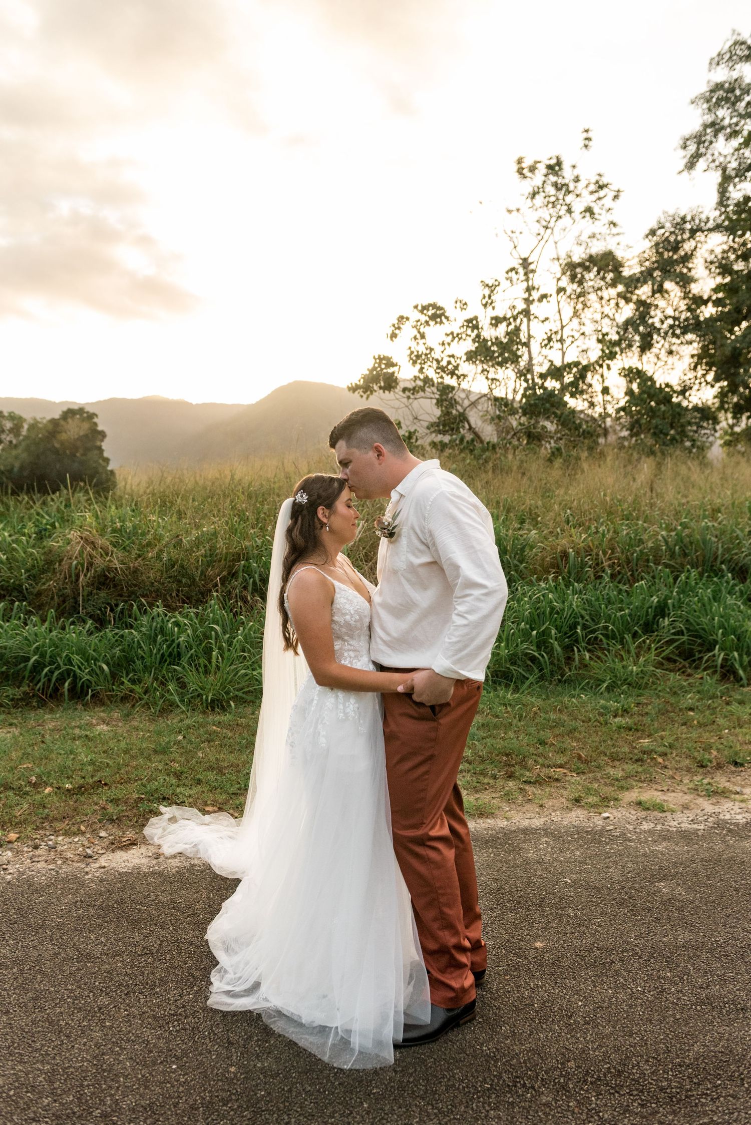 Bride and groom share a tender moment at golden sunset with mountain landscape behind.
