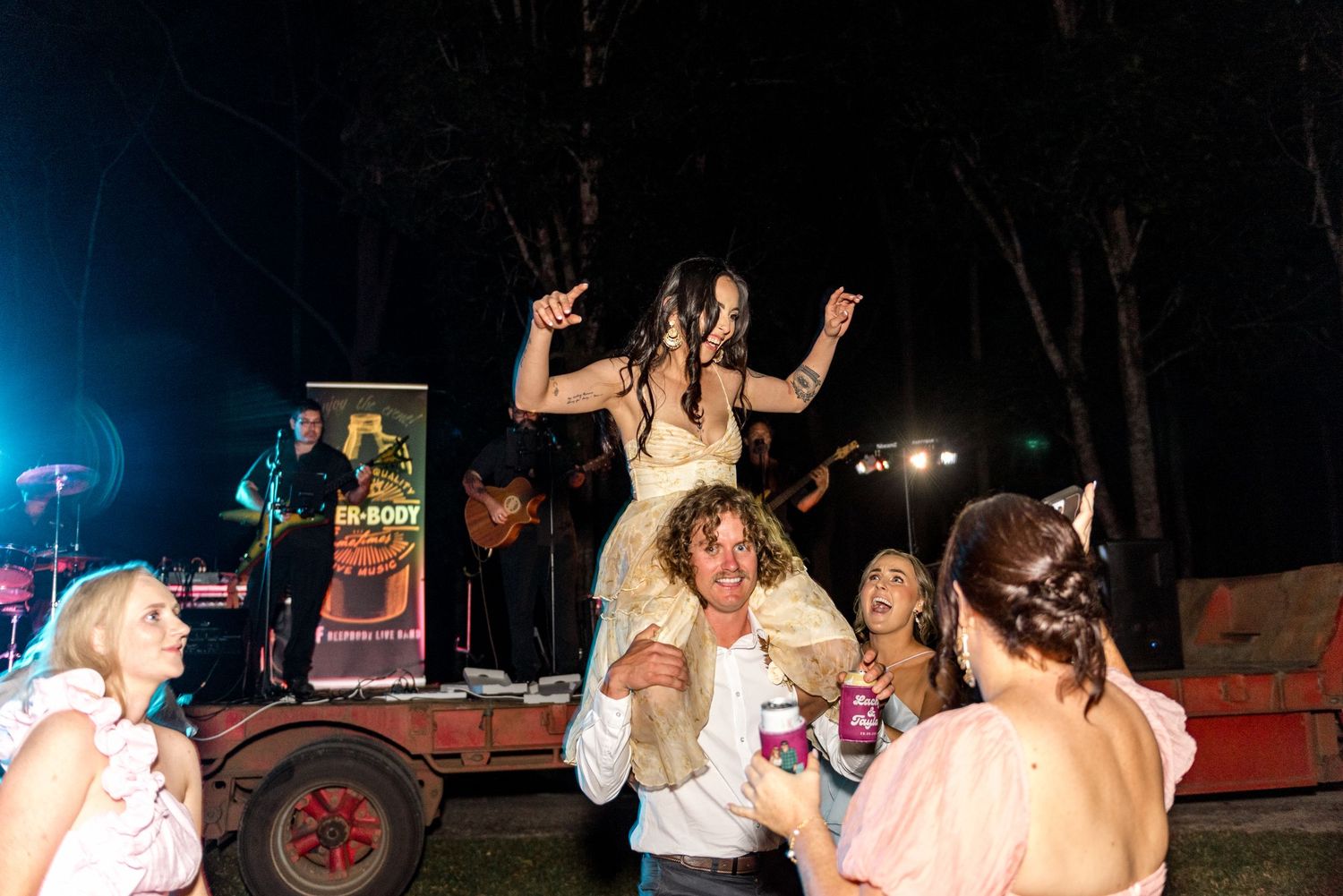 Bride lifted on shoulders dancing at lively outdoor nighttime wedding reception with band.