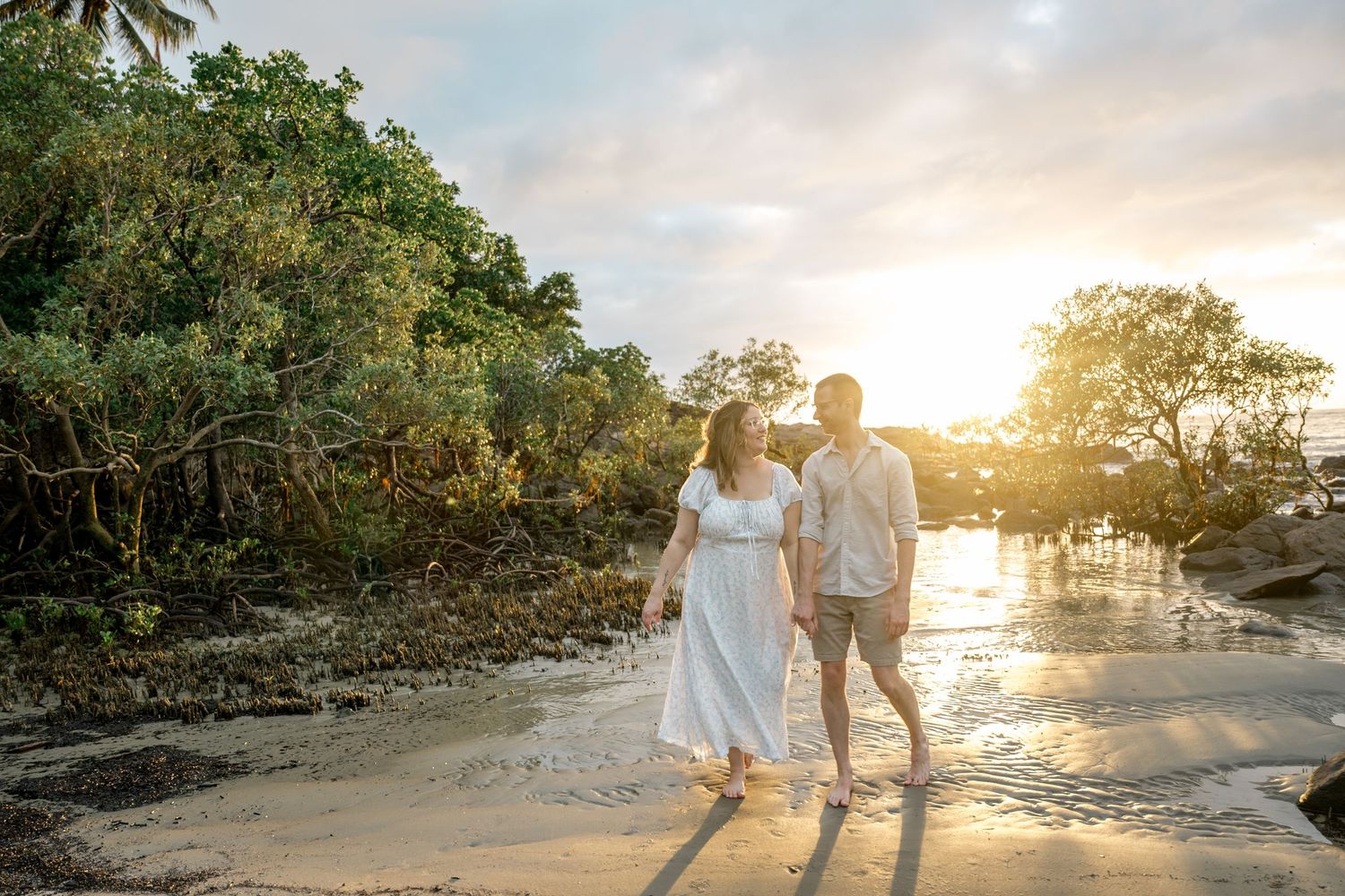 Couple walking hand in hand along tropical beach at sunset with lush green trees in background at Four Mile Beach, Port Douglas.