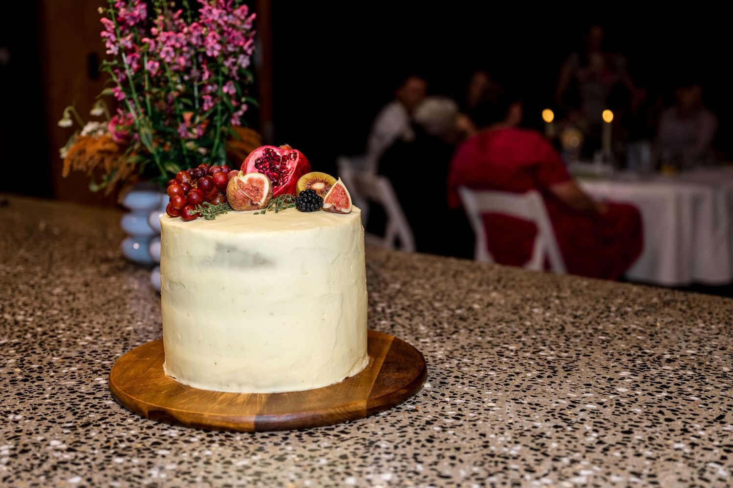 White frosted celebration cake topped with fresh fruit displayed on a granite countertop at an event.