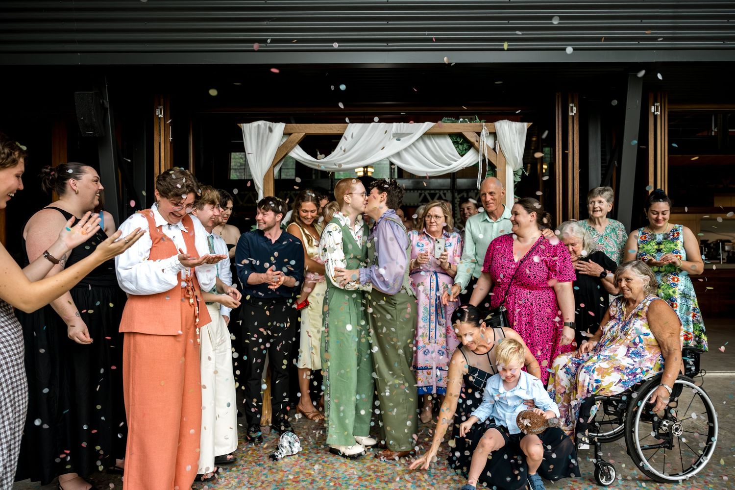 Wedding couple celebrates with family and friends throwing confetti outside rustic venue.