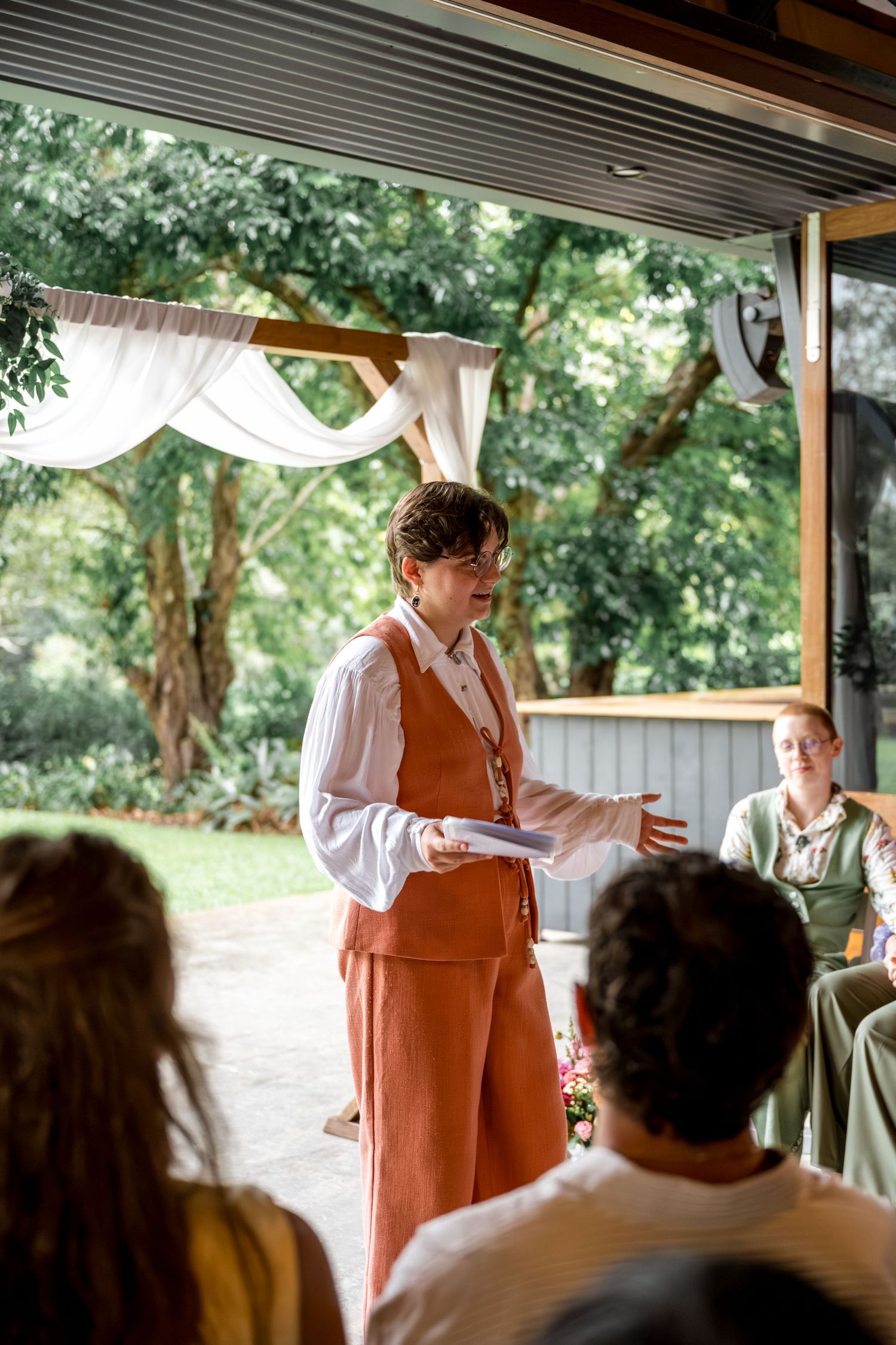 Wedding celebrant in orange linen vest speaks during an outdoor garden ceremony.