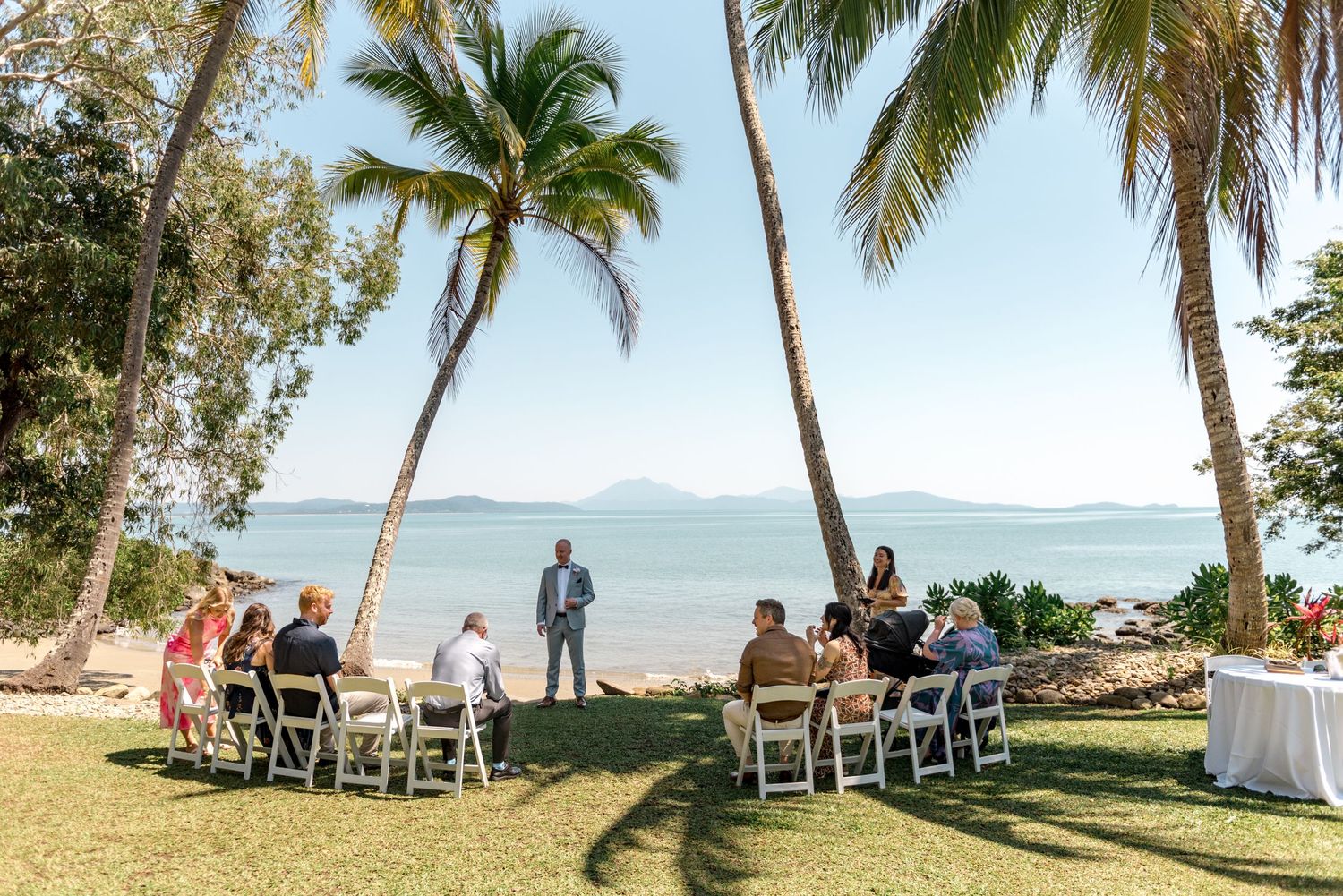 Intimate beachside wedding ceremony with guests seated on white chairs under tropical palm trees.